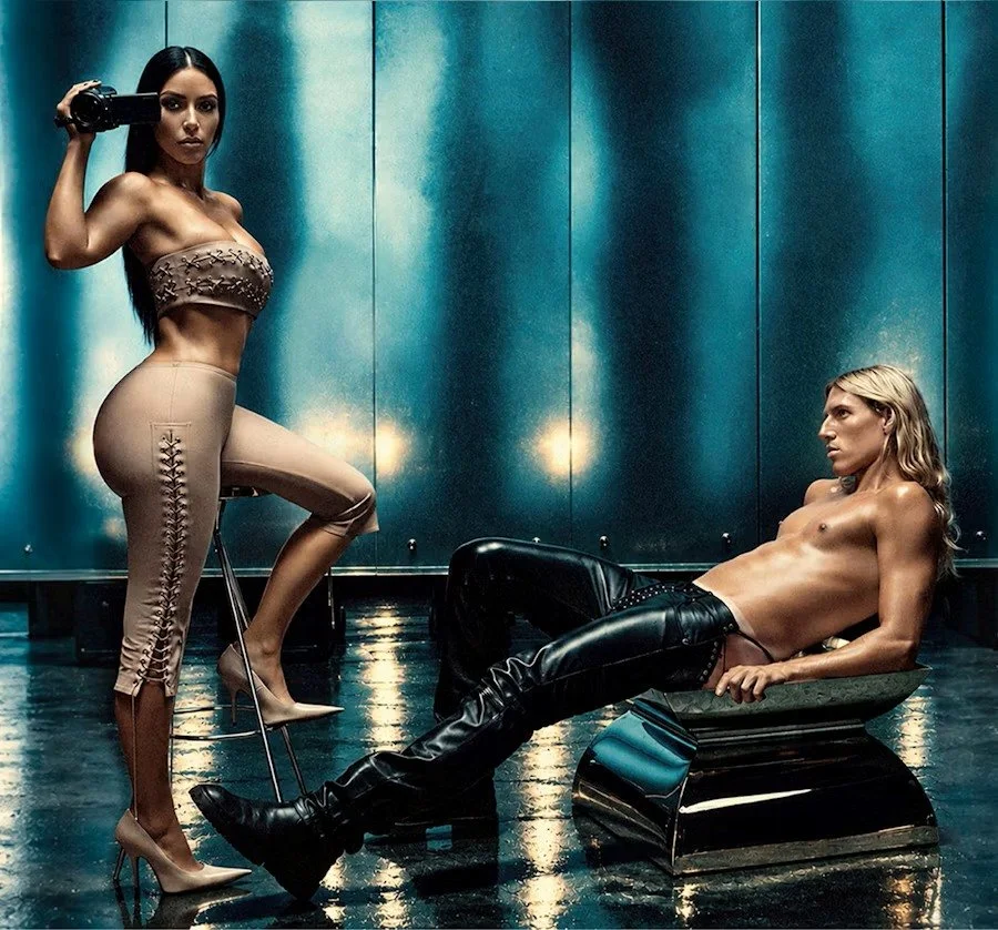 Kim Kardashian  standing with one leg raised,, wearing black leather pants and boots, is reclining on a metallic bench, looking at her in a dimly lit room with blue walls and reflective floor.