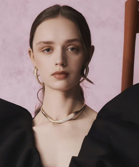 A young woman with brown hair in a low ponytail wearing gold hoop earrings, a gold necklace, and a black top. She is standing against a pink background.