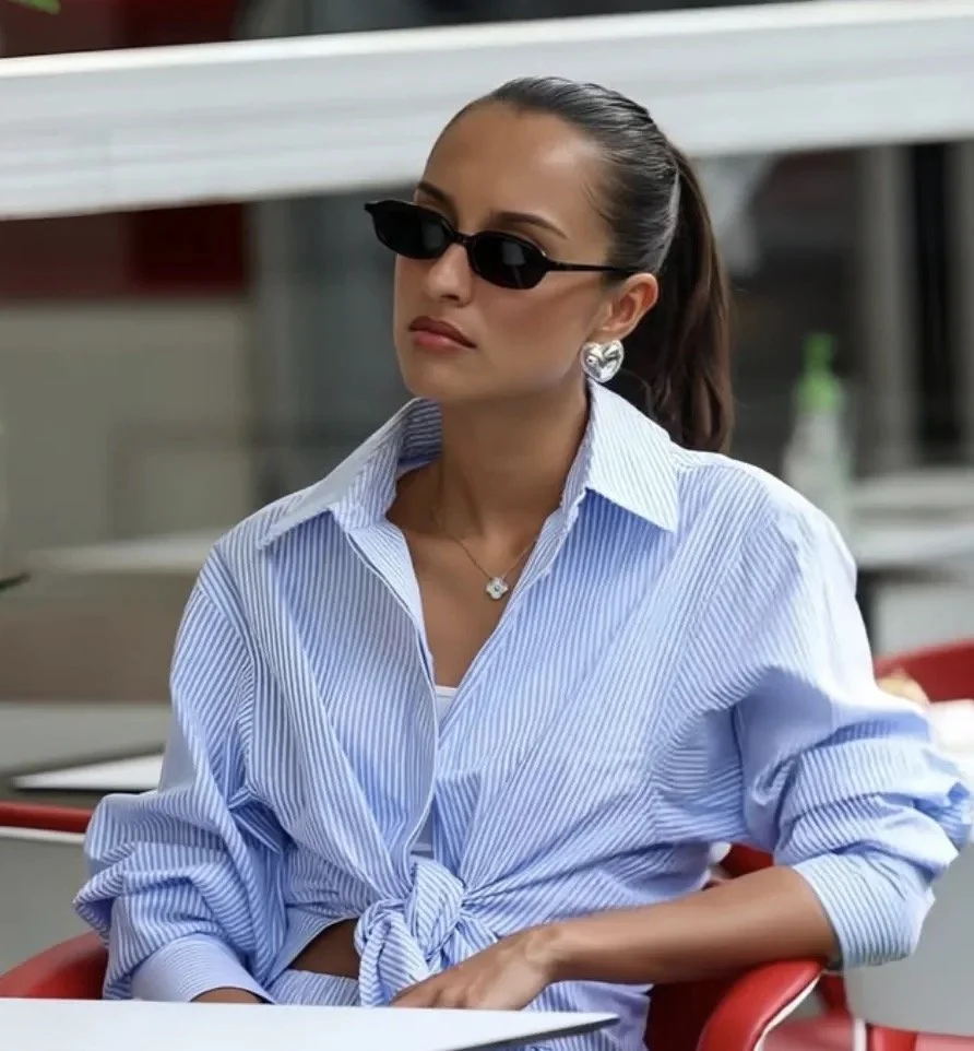 A woman wearing black sunglasses, a striped blue and white shirt tied at the waist, silver earrings, and a necklace, sitting outdoors.