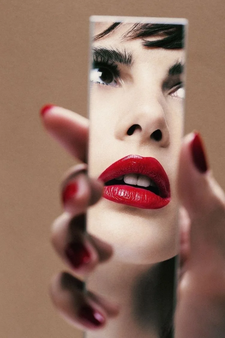 Close-up of a woman with bold red lipstick and dark hair, looking into a small mirror she is holding. The mirror reflects her face with a focus on her eye and lips.