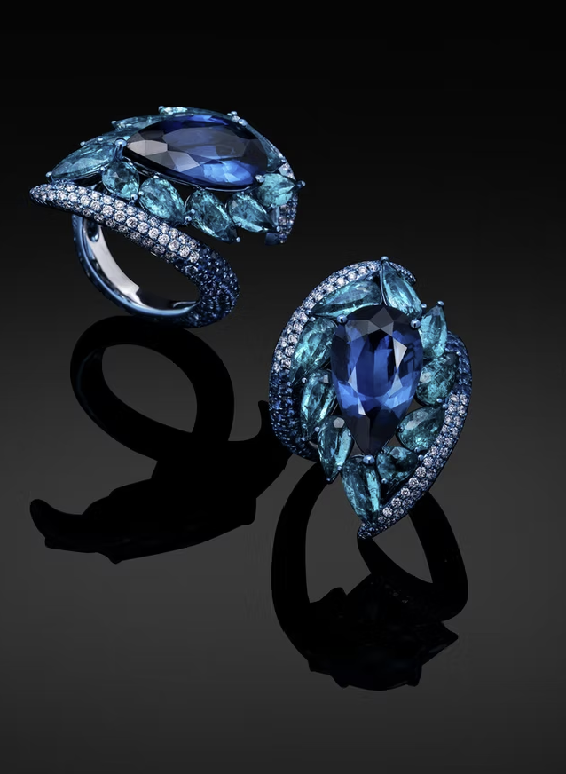 Blue gemstone rings with multiple smaller blue stones and diamonds, set in silver or white gold, displayed on a reflective black surface.