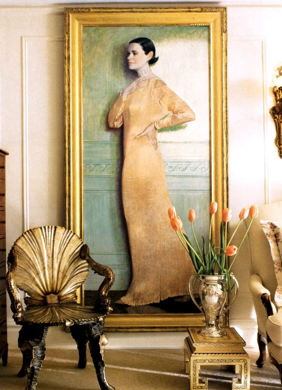 Decorative painting of a woman in a long beige dress with dark hair, displayed on a wall in a traditionally furnished room. The painting is framed in gold, and the room contains a gold-accented shell-shaped chair, a glass vase with pink tulips on a side table, and other elegant furnishings.