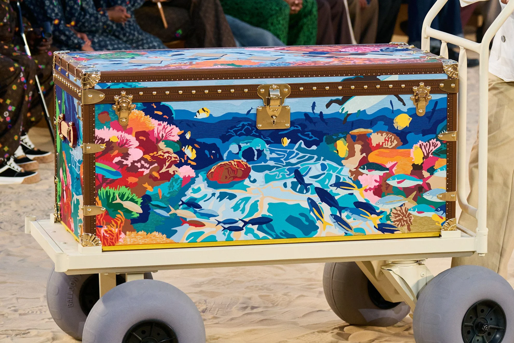 Decorative Louis Vuitton trunk with an ocean scene featuring colorful coral, fish, and underwater creatures, placed on a white wooden cart with gray wheels.