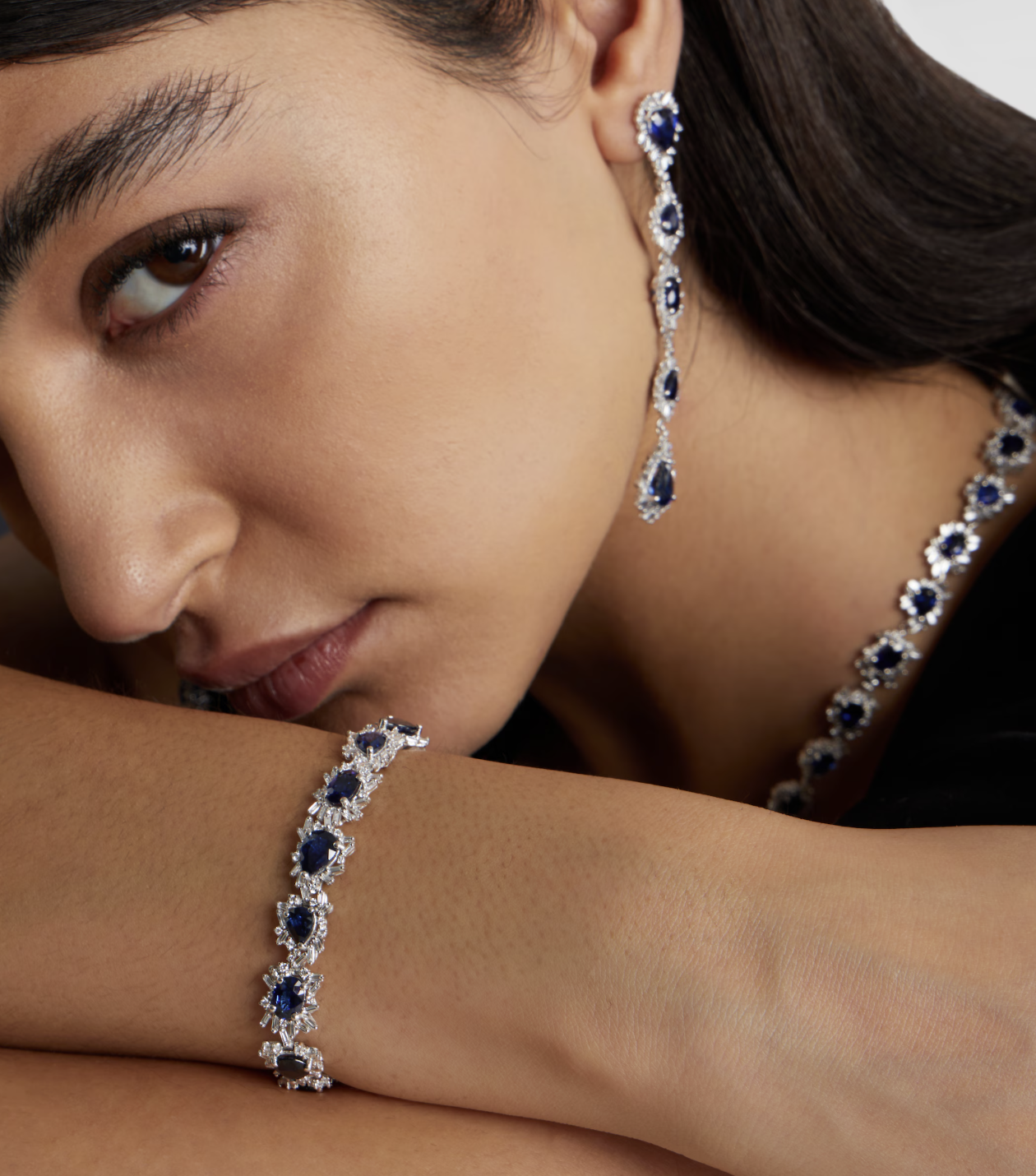 Close-up of a woman wearing diamond and sapphire jewelry, including earrings, a necklace, and a bracelet.