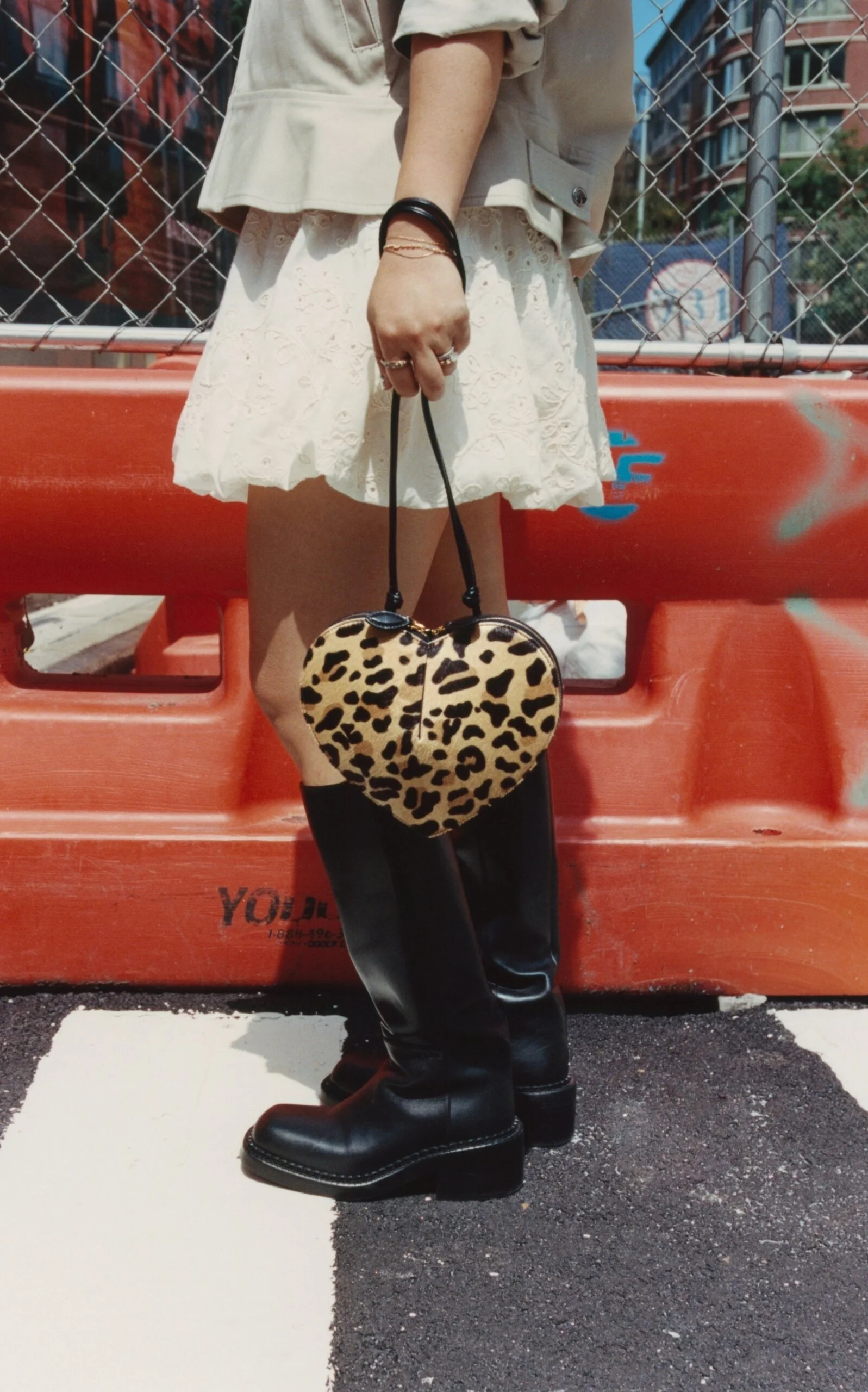 A person standing on the street holding a leopard-print heart-shaped handbag. The person is wearing black leather boots, white lace skirt, and a beige top with rolled-up sleeves. The background includes a red barrier, chain-link fence, and brick buildings.