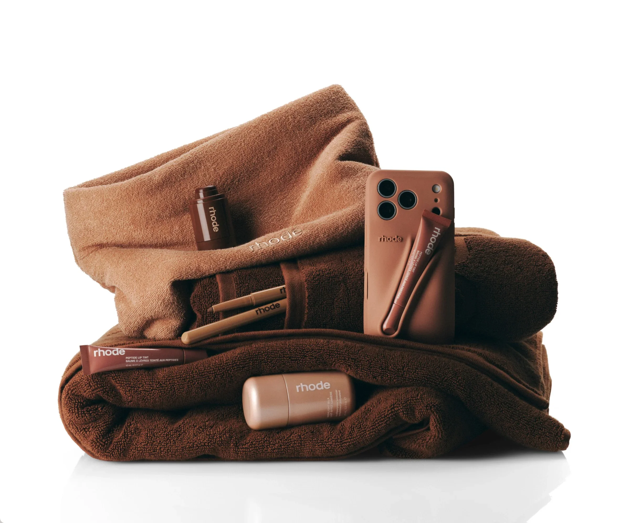 A collection of brown and beige skincare and beauty products, including lip tint, lip balm, and a facial mist, arranged on stacked brown towels.