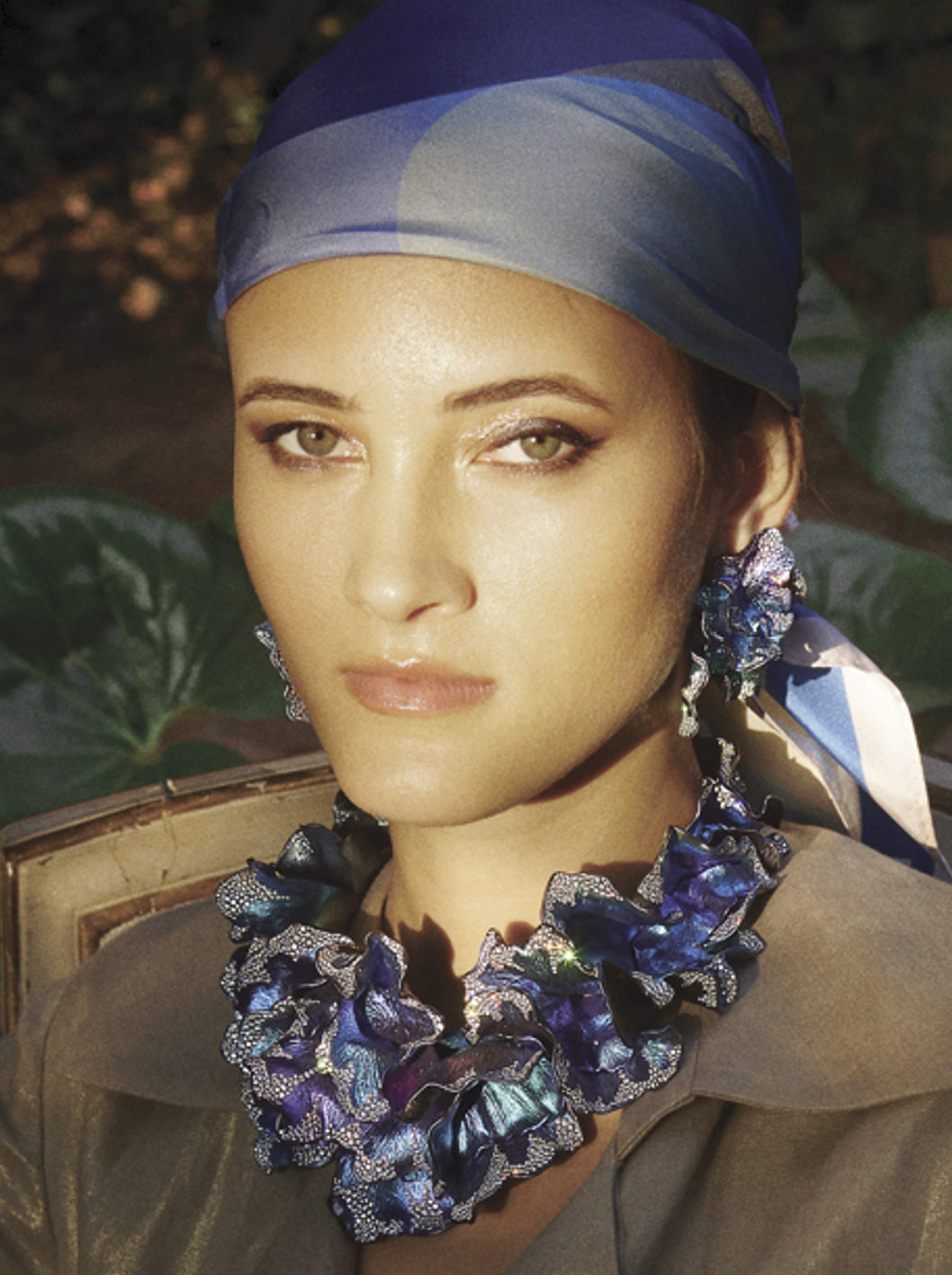 Woman with light skin and green eyes wearing a blue patterned headscarf, floral jewelry, and a green top, outdoors among greenery.