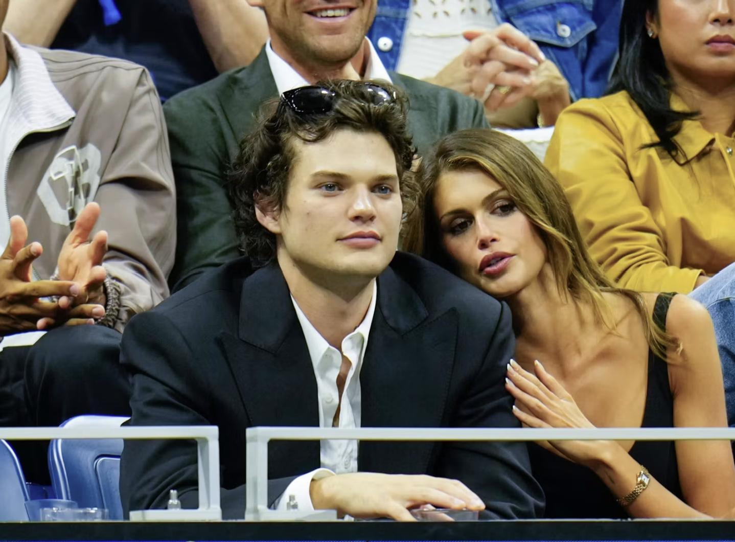A man and a woman sitting closely at a public event, with the woman resting her head on the man's shoulder. The man has curly brown hair, wears a dark suit, and sunglasses on his head. The woman has long wavy hair, wears a black tank top, and has a watch on her wrist.