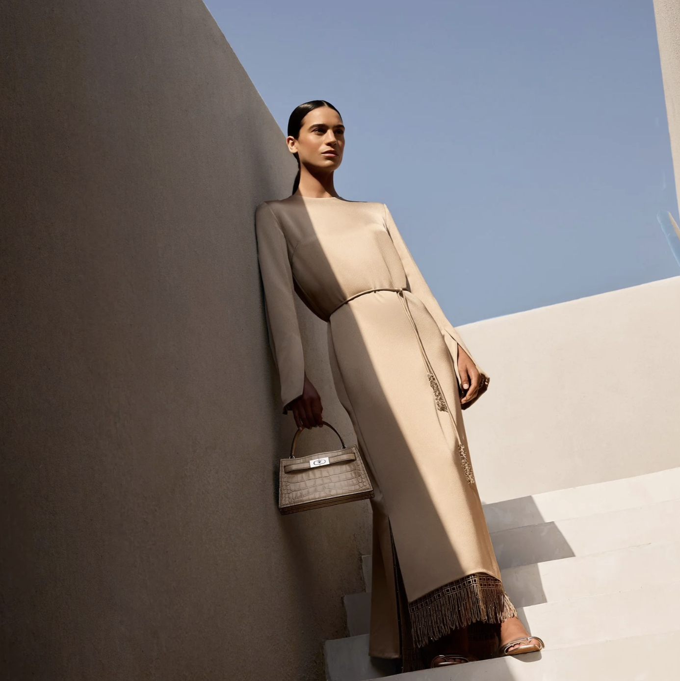 A woman dressed in beige standing on stairs against a minimalist modern backdrop under a clear blue sky, holding a small handbag.