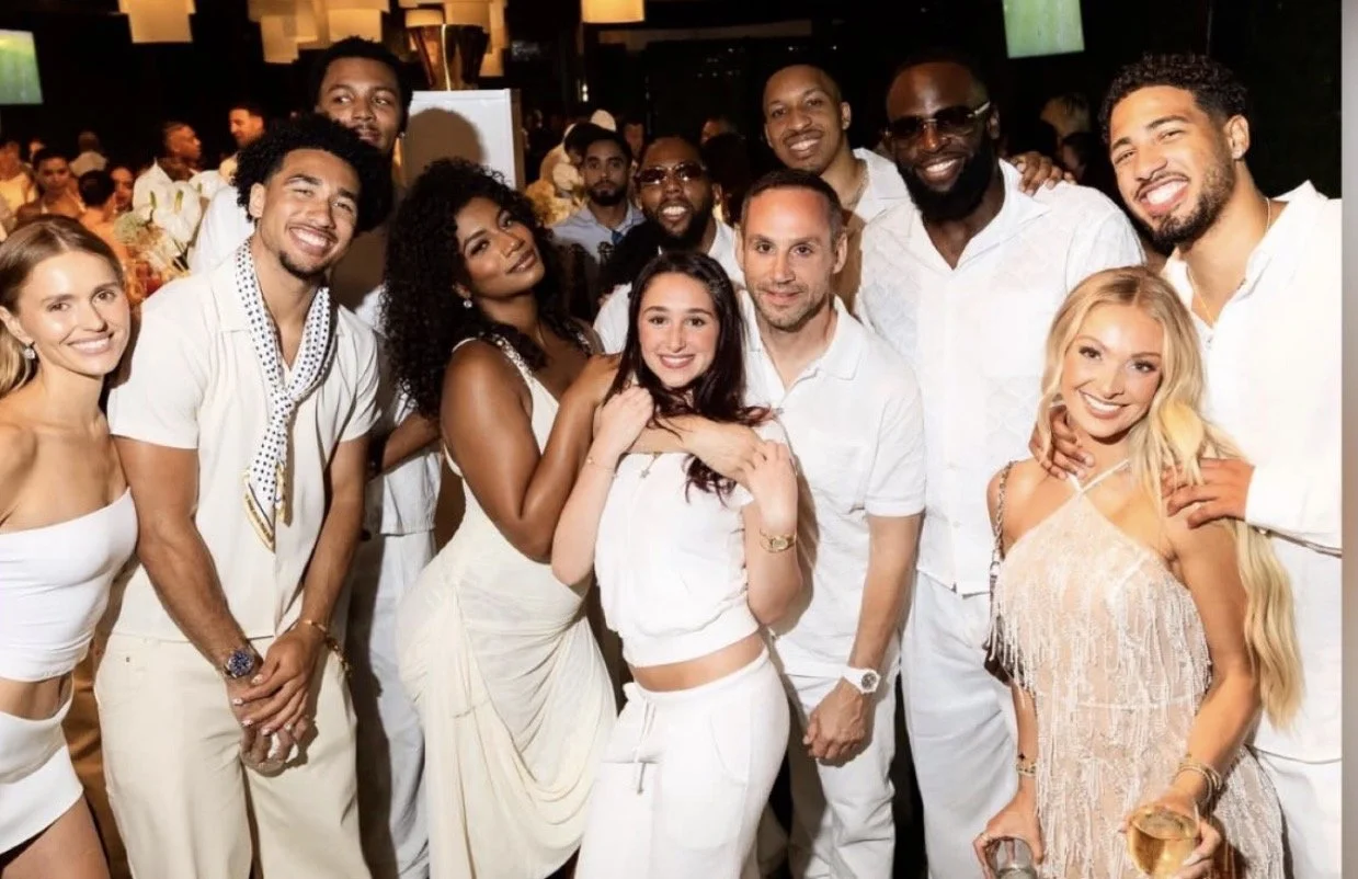 Group of people smiling and posing together at a social event, dressed in light-colored and white clothing, with a background of more guests and decorations.