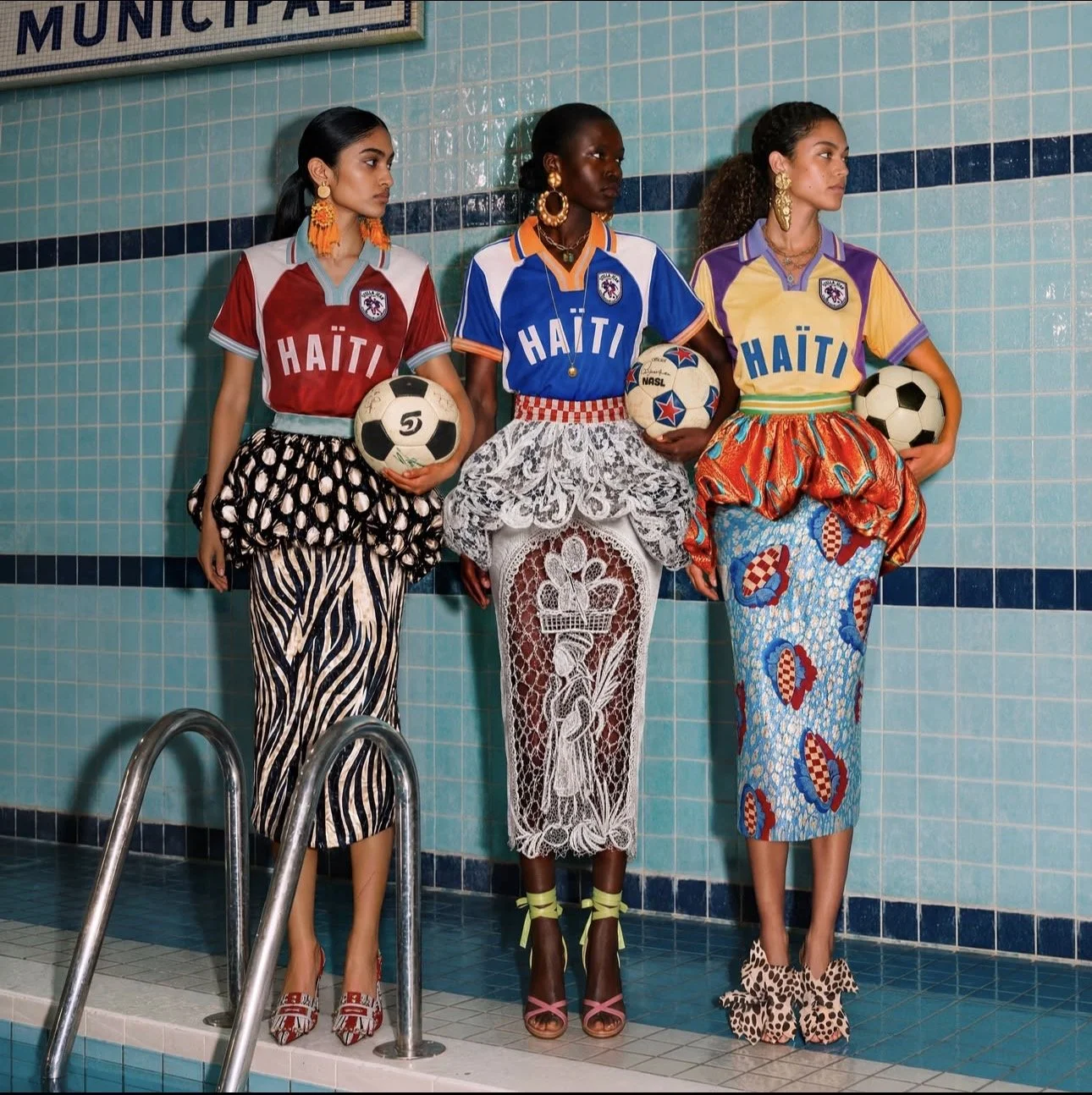 Three women standing by a swimming pool, holding soccer balls, wearing colorful patterned skirts and T-shirts that say 'Haiti' with portraits on the shirts, against a blue tiled wall, with a sign partially visible that reads 'Municipal'.
