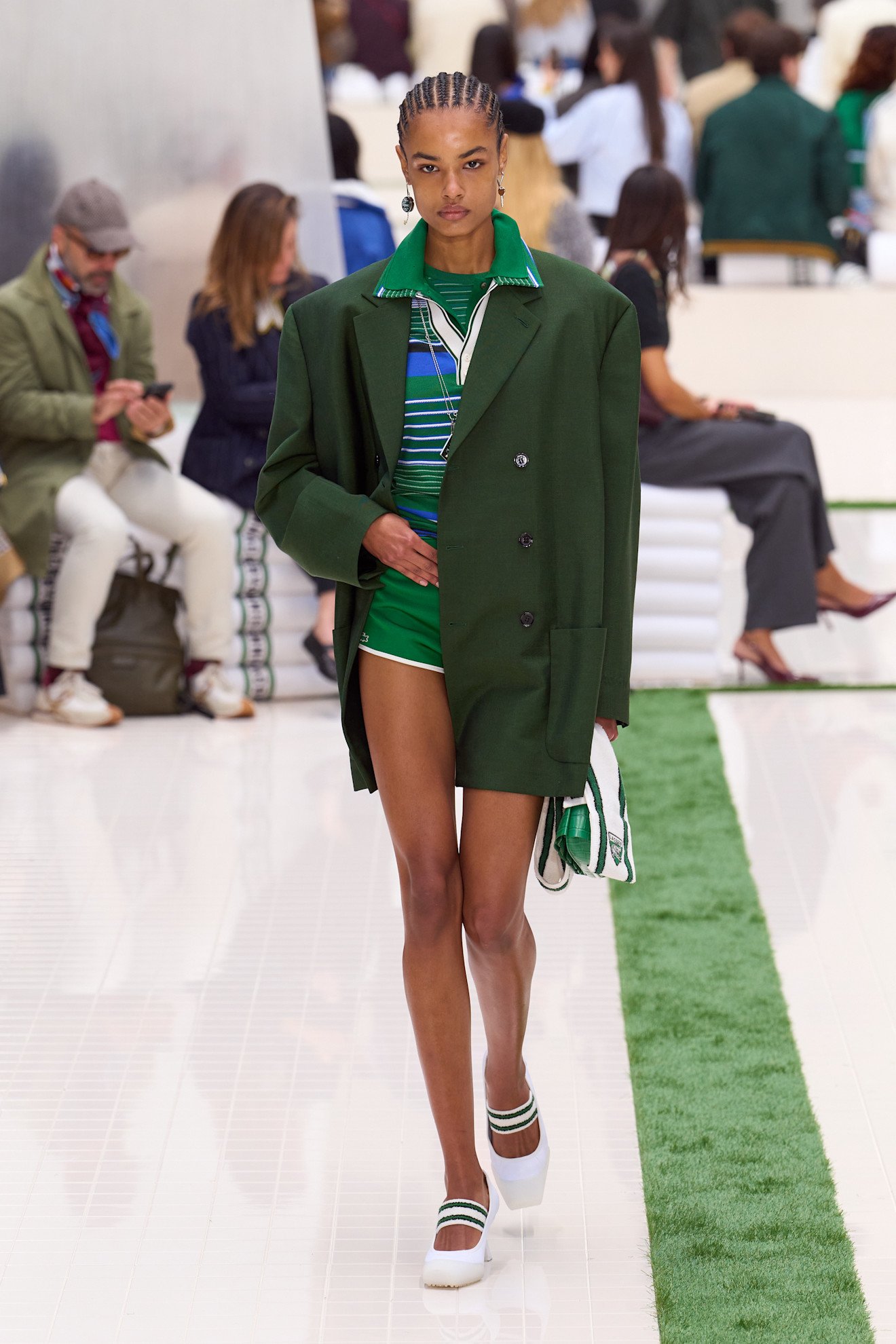 Fashion model walking on runway wearing a green oversized coat, green striped shorts, and white shoes with green stripes, carrying a small bag, with an audience in the background.