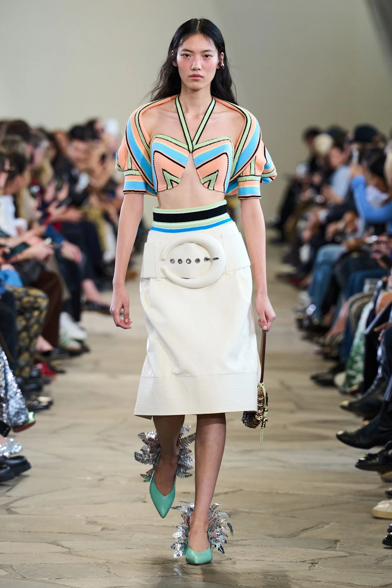 A fashion model walking down the runway during a fashion show, wearing a colorful, geometric patterned top, a white skirt with a large circular belt buckle, turquoise high heels with metallic feather-like accents, and carrying a small handbag.