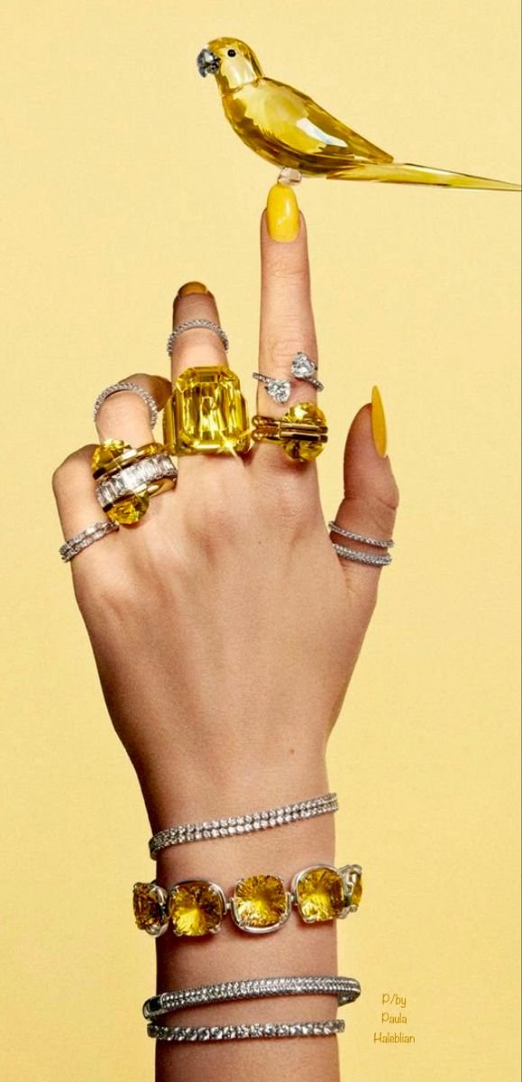 A hand with yellow-painted nails is adorned with multiple rings, bracelets, and watches. A yellow bird is perched on the index finger.