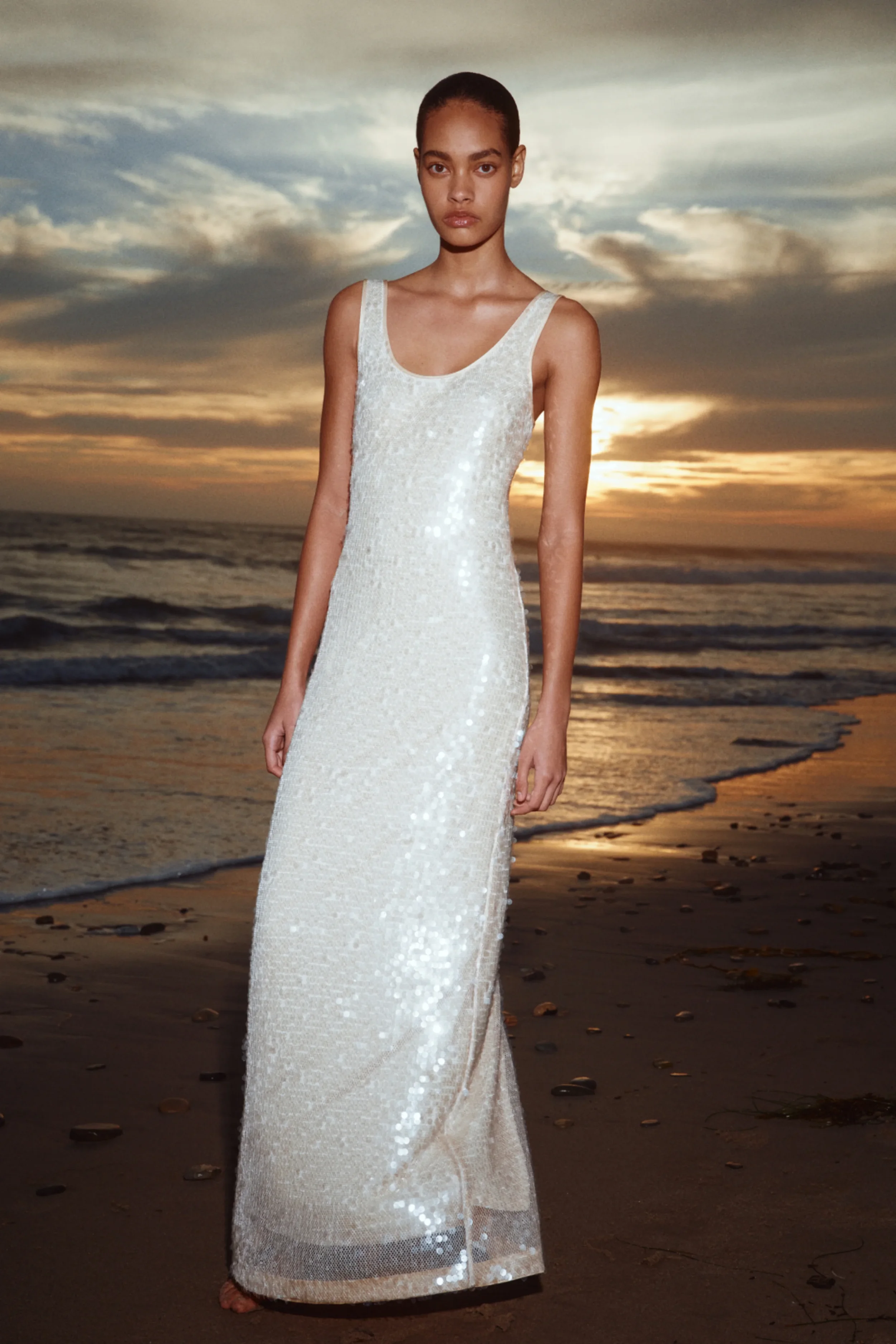 A woman in a shiny, white sleeveless dress stands barefoot on a beach at sunset, with a cloudy sky and ocean in the background.