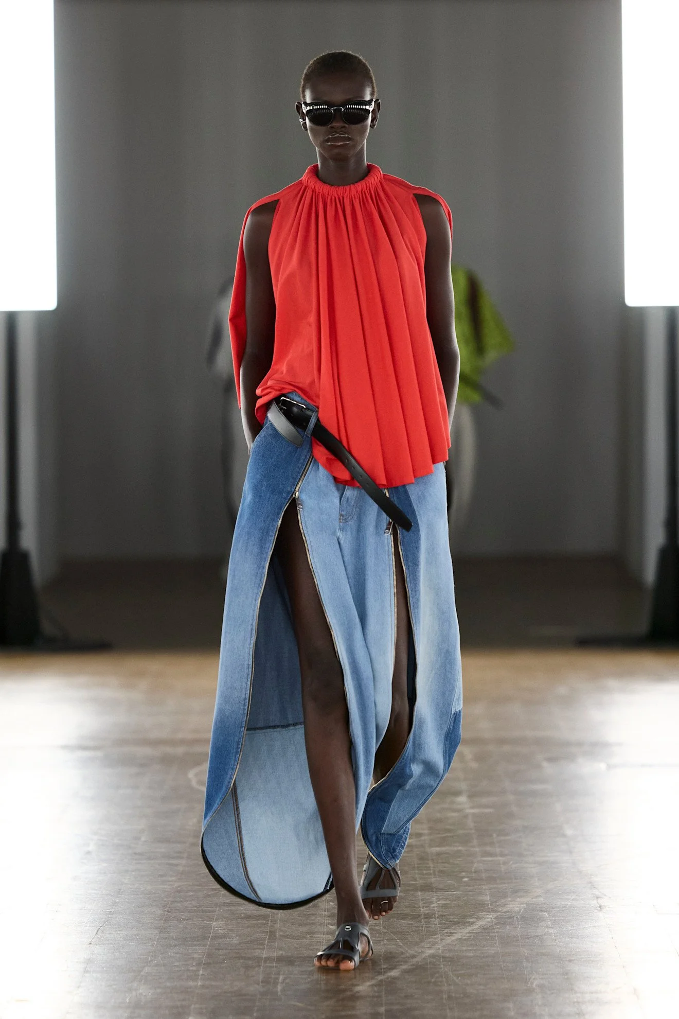 Model walking runway wearing a red pleated sleeveless top with shoulder cutouts, a long denim skirt with zipper details and a thigh slit, paired with sandals, sunglasses, and a belt with a pouch, in a fashion show setting.