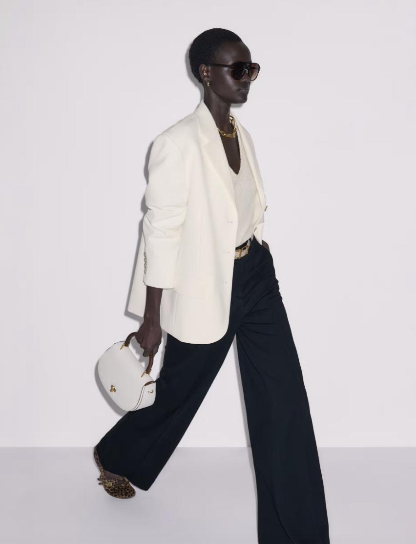 A stylish woman with dark skin wearing a cream blazer, white top, black wide-leg pants, dark sunglasses, and leopard print shoes. She holds a white handbag with gold details and wears gold jewelry, standing against a plain white background.