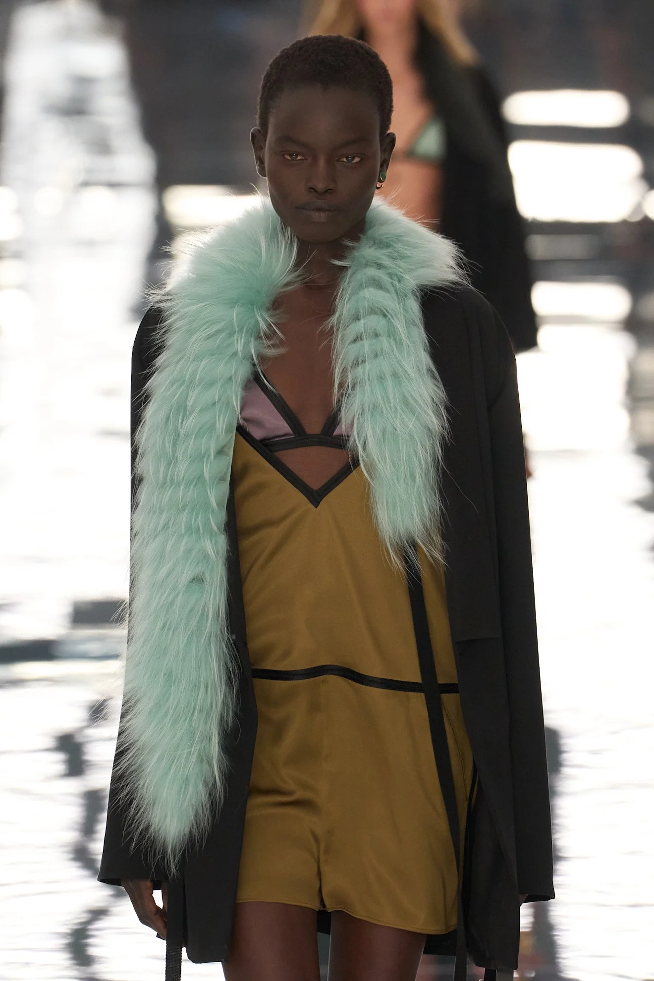 A female model walking on a runway wearing a black coat with light green faux fur collar, a yellow dress with black accents, and earrings, with blurred background and bright reflections.