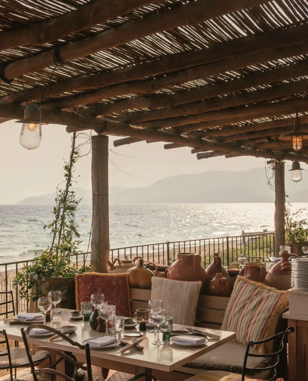 An outdoor dining area overlooking the ocean with a table set with glassware, plates, and silverware under a rustic wooden roof. There are chairs with covered cushions and decorative vases on the table, with large pottery vases along the railing, and mountains visible in the background during sunset.