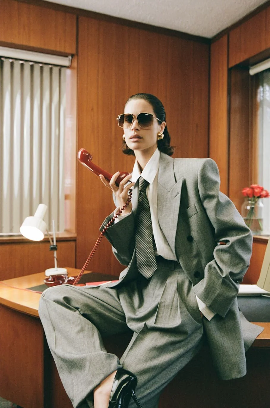 A woman in a gray suit, white shirt, and tie sitting on a desk, holding a red landline phone, wearing sunglasses and gold earrings, in an office with wood-paneled walls and a window with blinds.