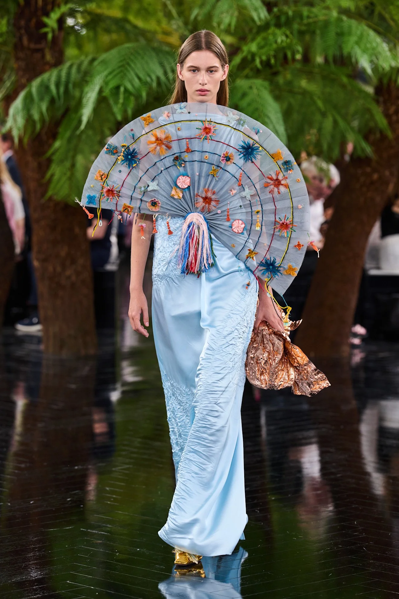 Fashion model walking on runway holding a decorated parasol, wearing a light blue silky gown with floral embroidery, gold shoes, and gold-colored accessories, background with lush green trees, other models and audience.