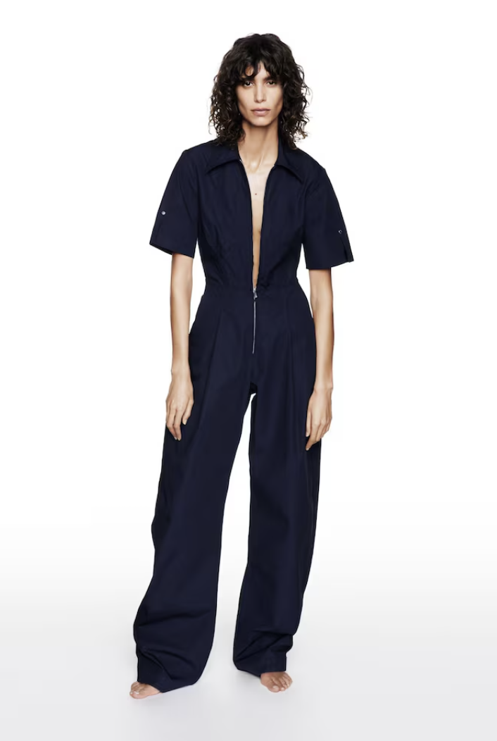 A woman with curly dark hair wearing a navy blue jumpsuit with short sleeves and a zipper front, standing barefoot against a white background.