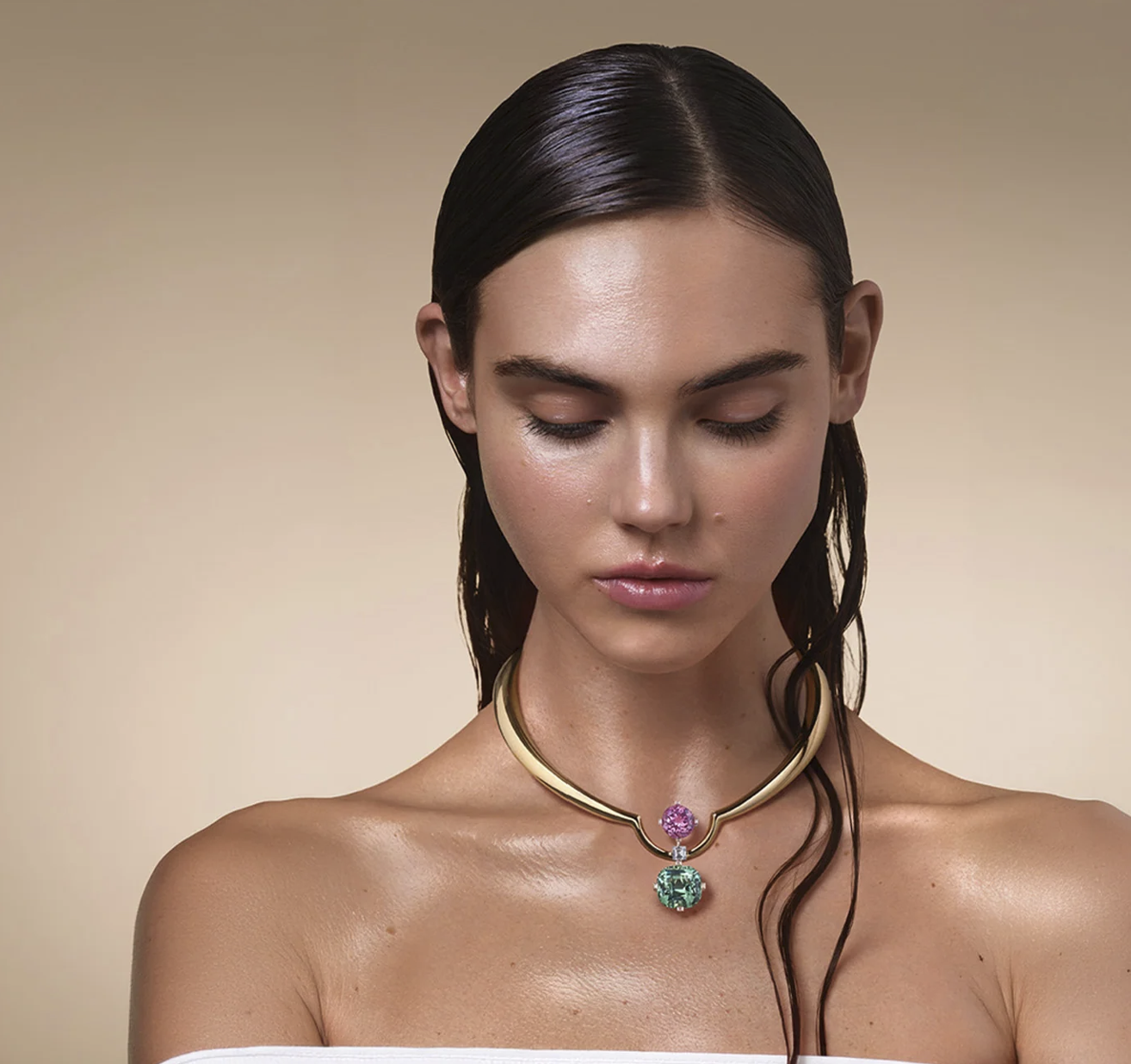 A woman with dark, slicked back hair wearing a gold necklace with two large colorful gemstones, one pink and one green, against a plain background.