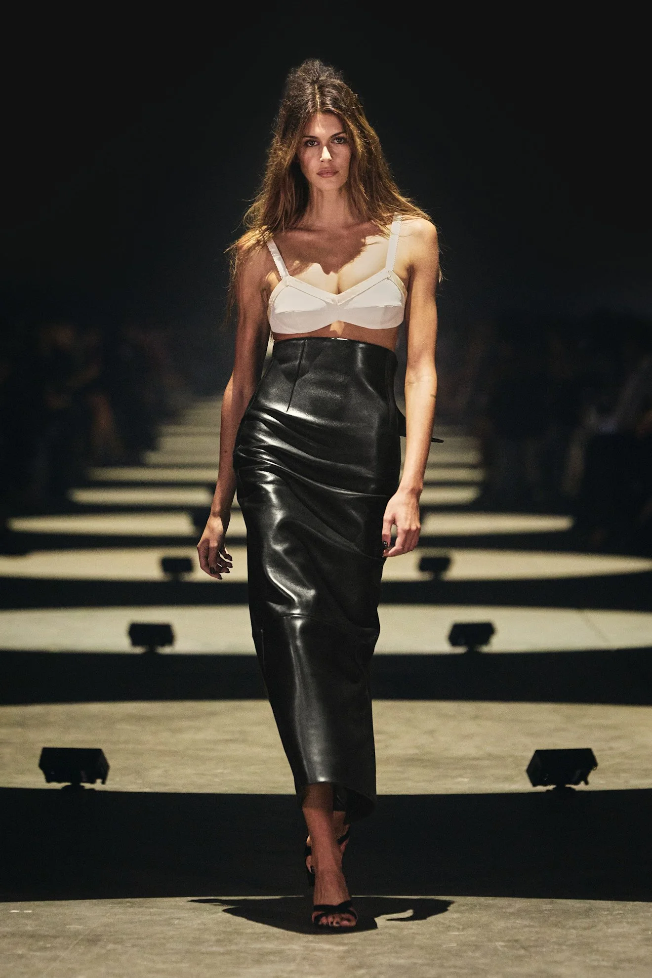 Kaia Gerber  walking down a runway, wearing a white crop top and a high-waisted black leather skirt, with a dark background and stage lighting.