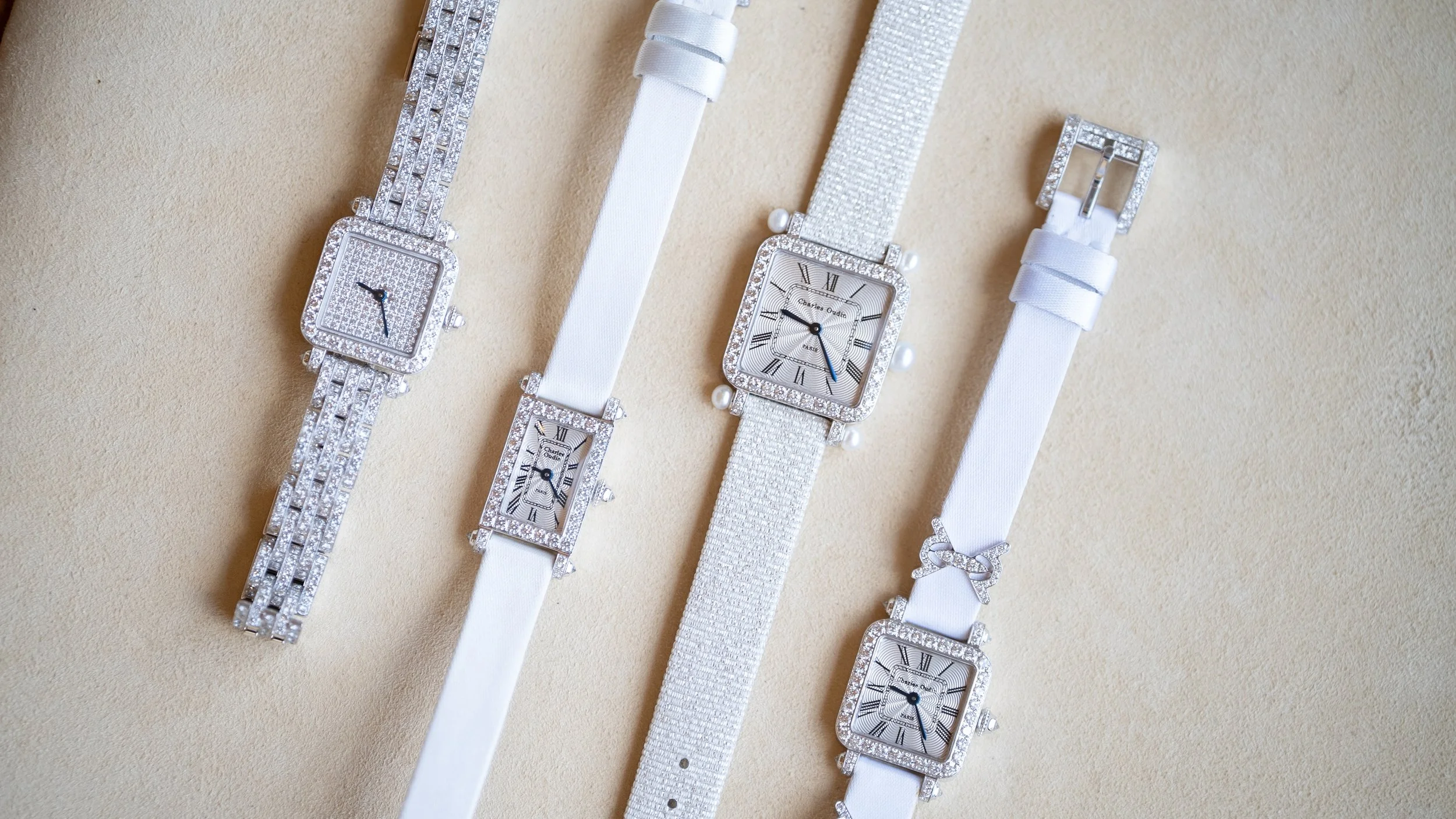 Four luxury women's wristwatches with white bands and square faces, decorated with crystals, laid out on a beige surface.