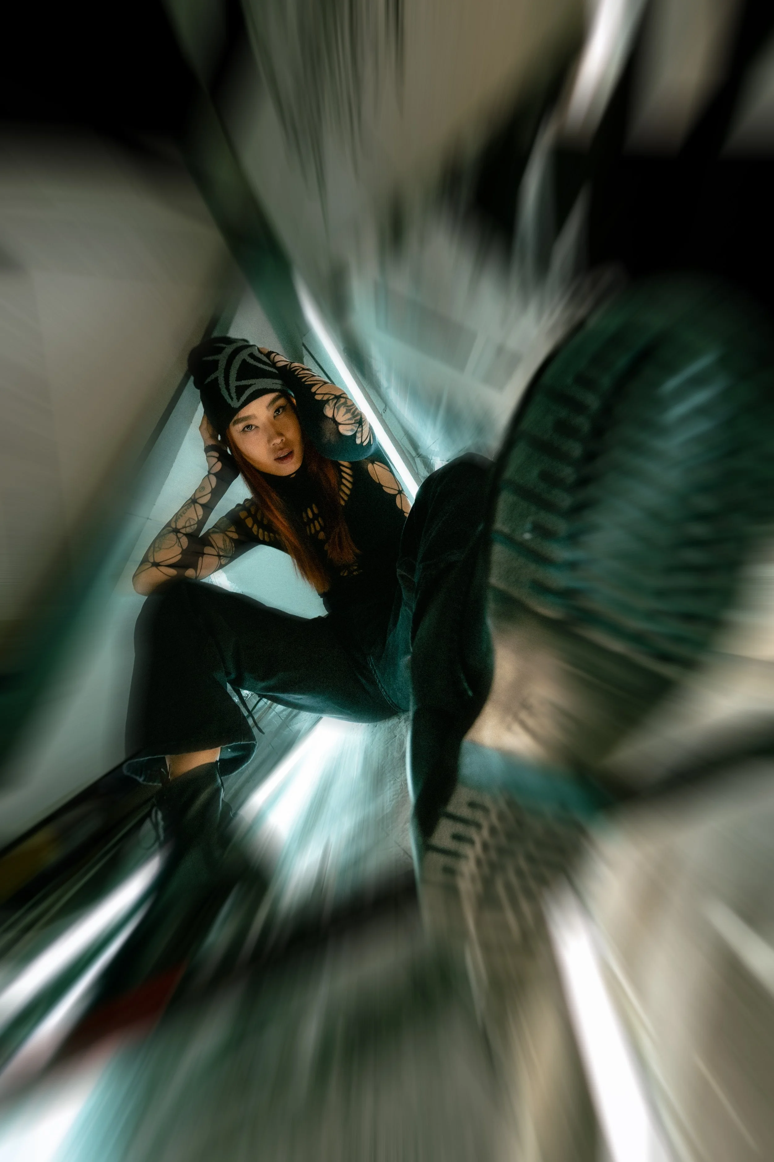 Junge Frau in schwarzer Kleidung mit durchsichtigen, netzartigen Ärmeln, die auf dem Boden in einem engen Raum sitzt und auf die Kamera blickt.