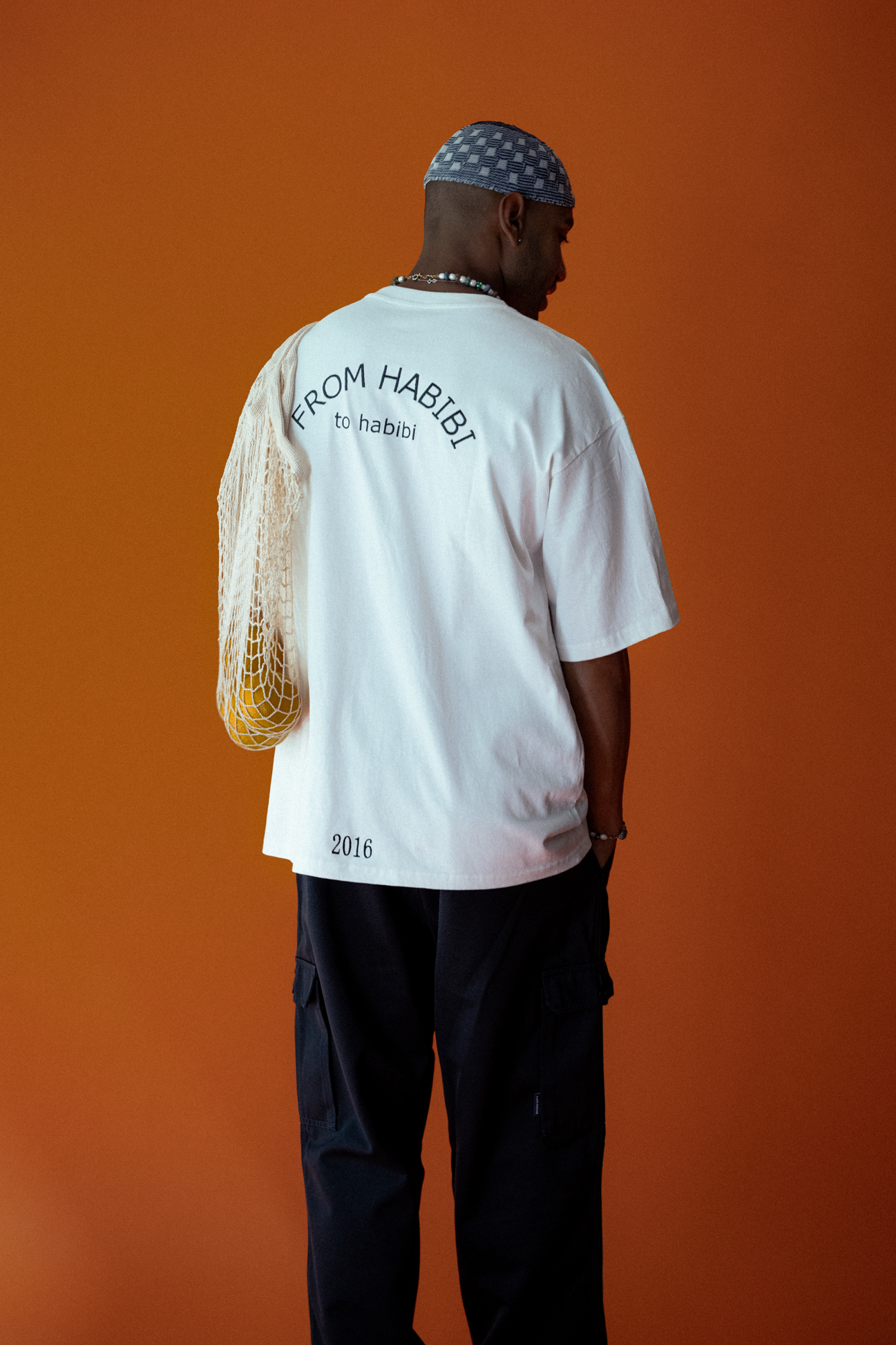 Ein junger Mann trägt ein weißes T-Shirt mit der Aufschrift 'FROM HABIBI to habibi' und die Jahreszahl 2016, dazu schwarze Hosen, mit einem blauen Musterndas Kopftuch und hält eine Netztasche. Der Hintergrund ist orange.