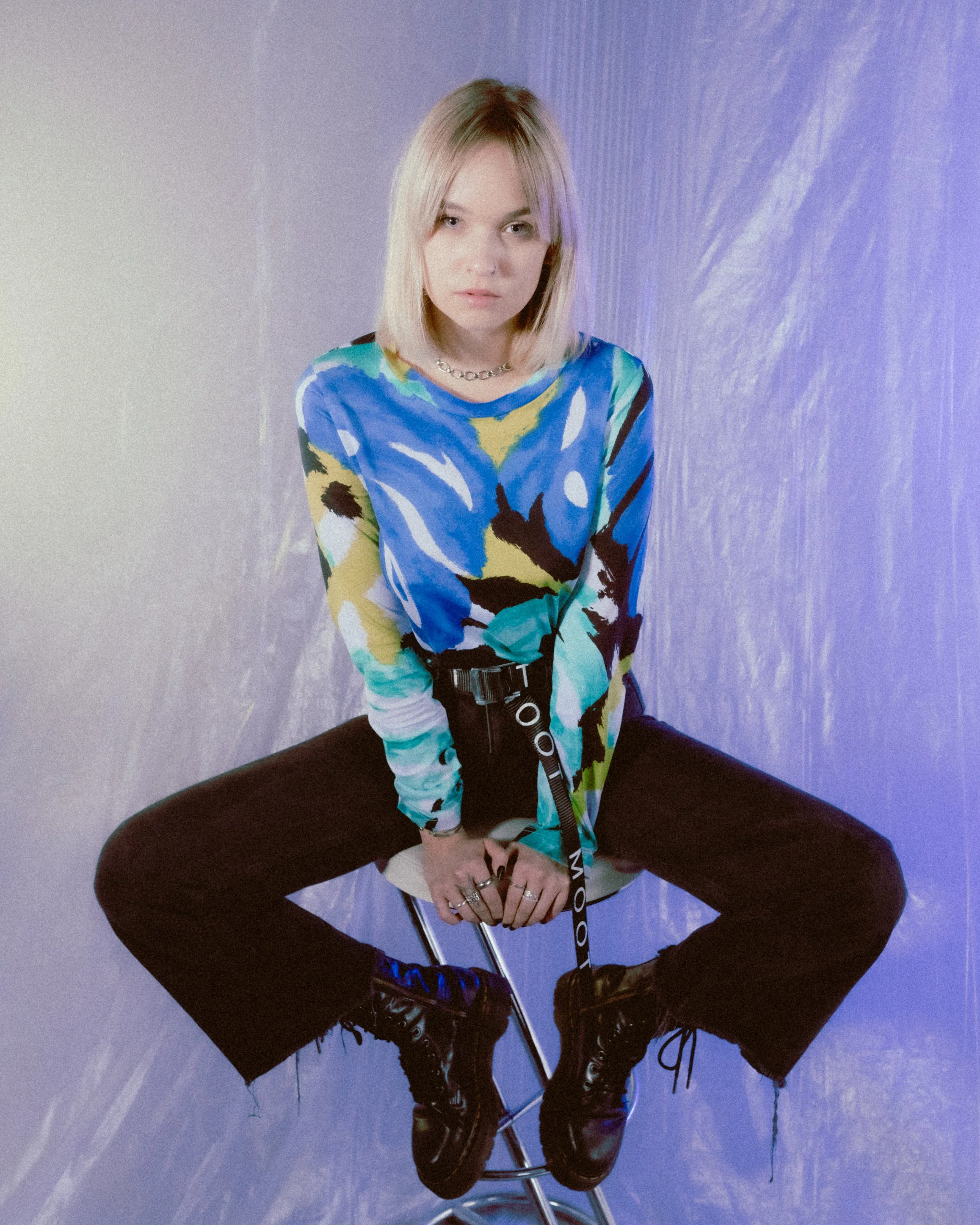 Junge Frau sitzt auf einem Stuhl, trägt eine bunte, abstrakte Langarmbluse, schwarze Hosen und hohe schwarze Schuhe, vor einem lilafarbenen Hintergrund.