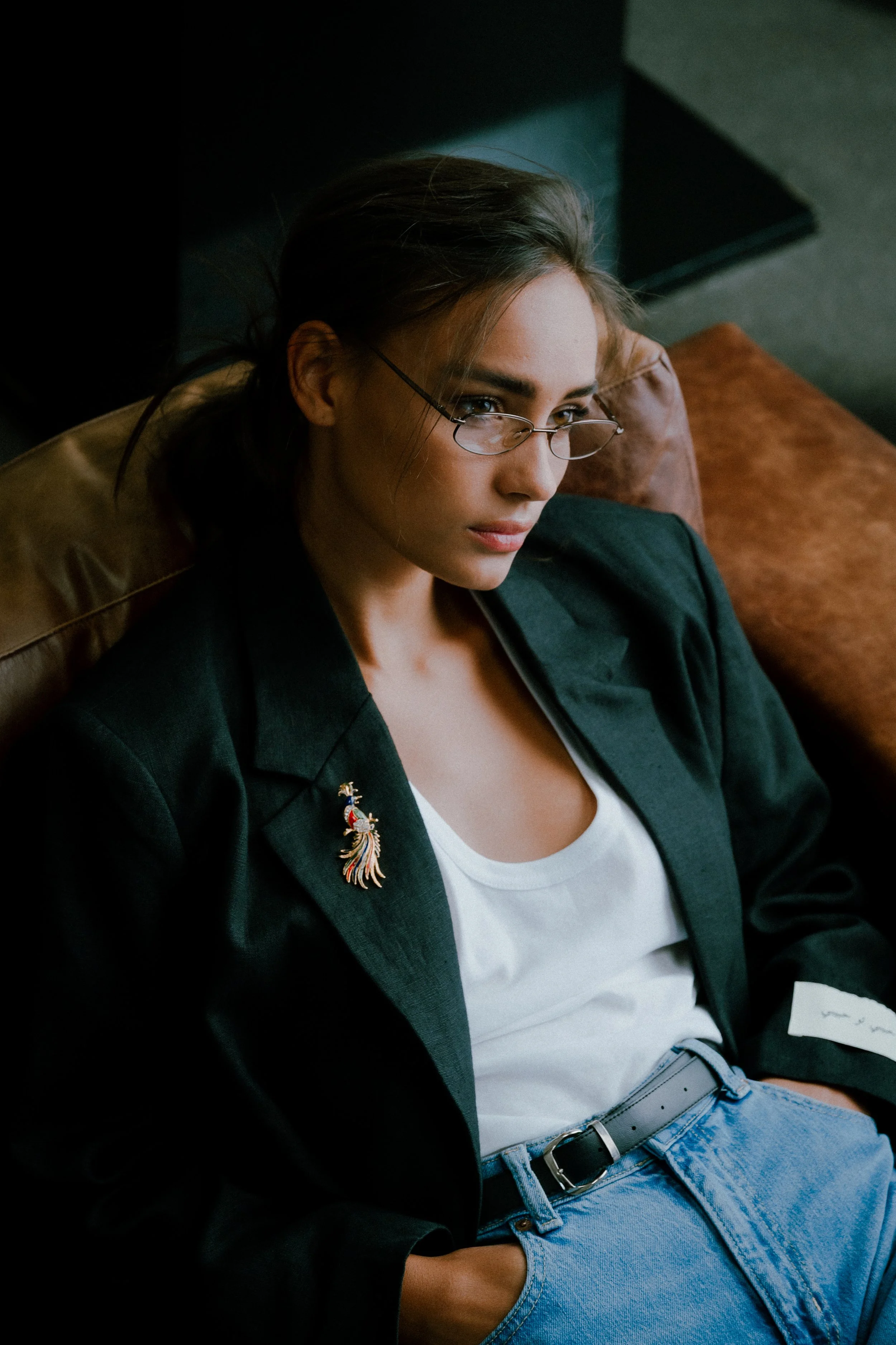 Junge Frau mit Brille sitzt nachdenklich auf einem Sofa, trägt eine schwarze Jacke, weiße T-Shirt, und eine Brosche an der Jacke.