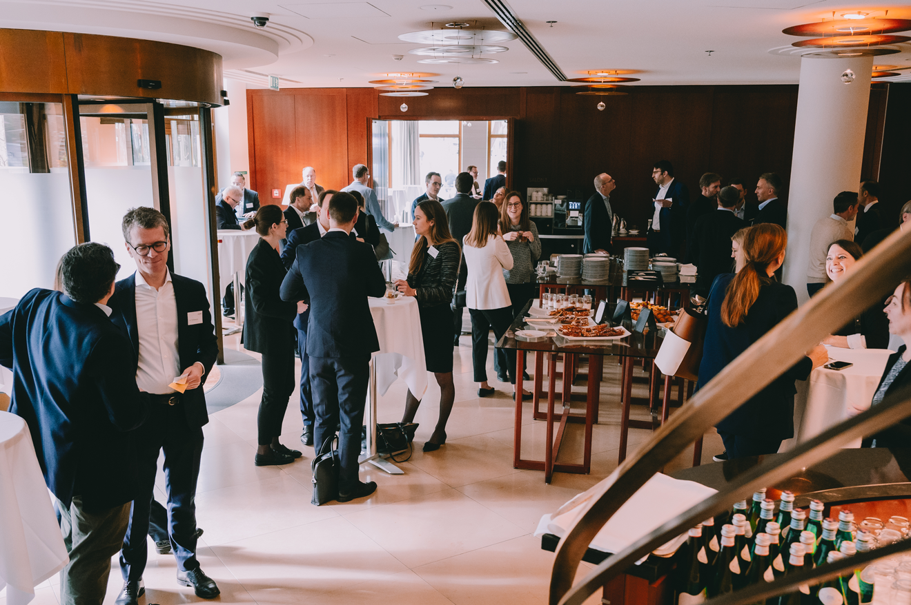 Geschäftsleute bei einer Netzwerken-Event in einem Hotelrestaurant, mit Stehtischen, Buffet mit Essen und Kaffee. Viele Personen in formeller Kleidung, einige mit Namensschildern.