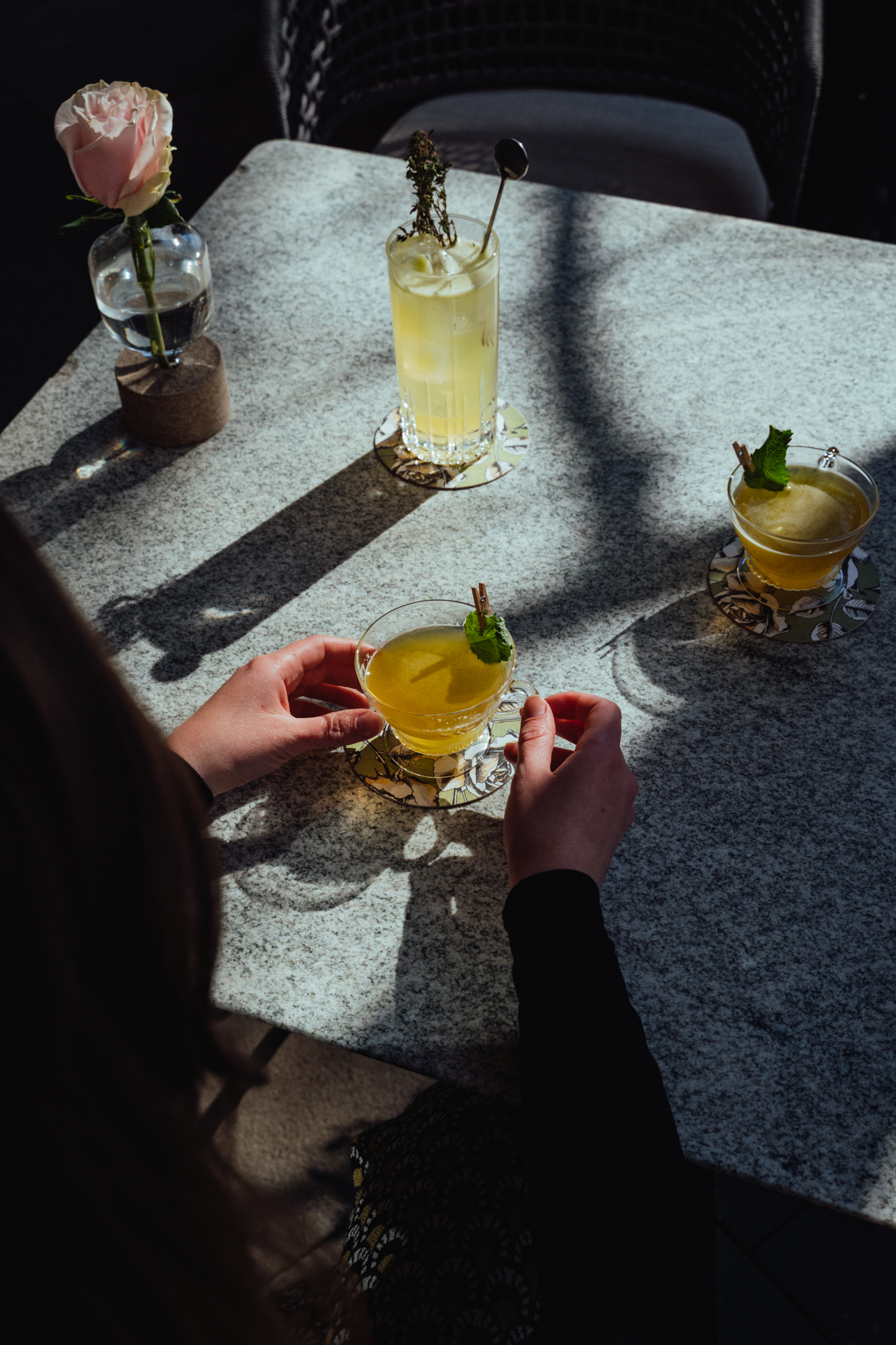 Person hält eine gelbe Cocktail mit Minzblatt auf einer grauen Marmor-Tischplatte. Im Hintergrund stehen zwei weitere Cocktails auf dekorativen Untersetzern, daneben eine kleine Vase mit einer rosa Rose.