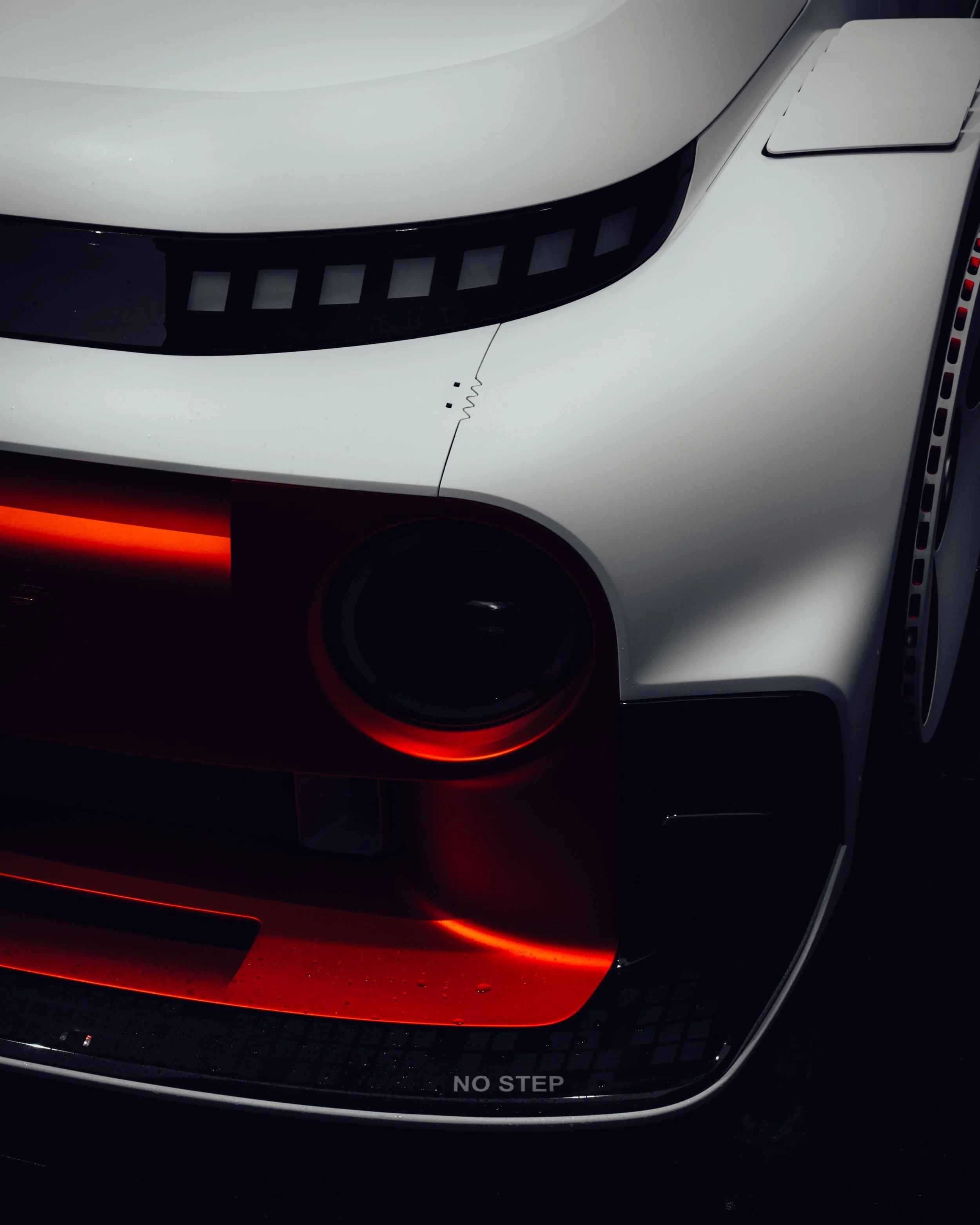 Nahaufnahme der Front eines weißen Autos mit schwarzen Akzenten und roter Beleuchtung, der Scheinwerfer ist sichtbar, der Aufkleber "NO STEP" ist am unteren Rand sichtbar.