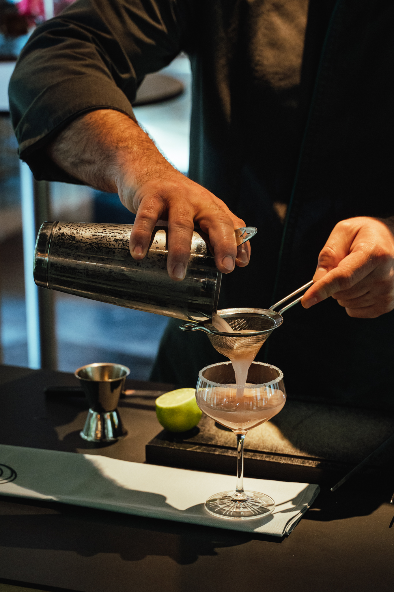 Ein Barkeeper siebt einen Cocktail in ein stilvolles Glas, während er ihn mit einem Cocktail-Shaker zubereitet. Im Hintergrund sind Limettenscheiben zu sehen.