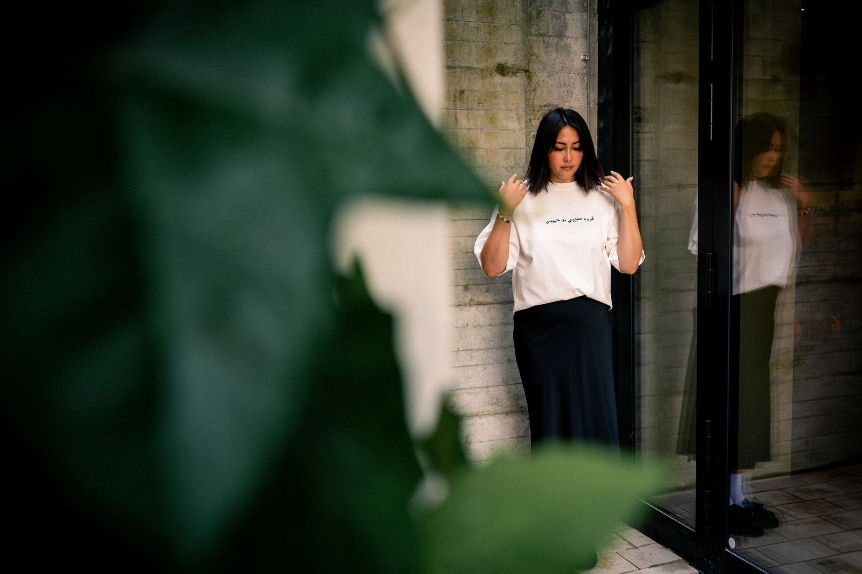 Junge Frau steht an einer Wand neben einer Glas Tür, sieht nach unten, trägt ein weißes T-Shirt mit arabischem Aufdruck und einen schwarzen Rock. Ihr Spiegelbild ist im Glas sichtbar, im Vordergrund sind große grüne Blätter unscharf.