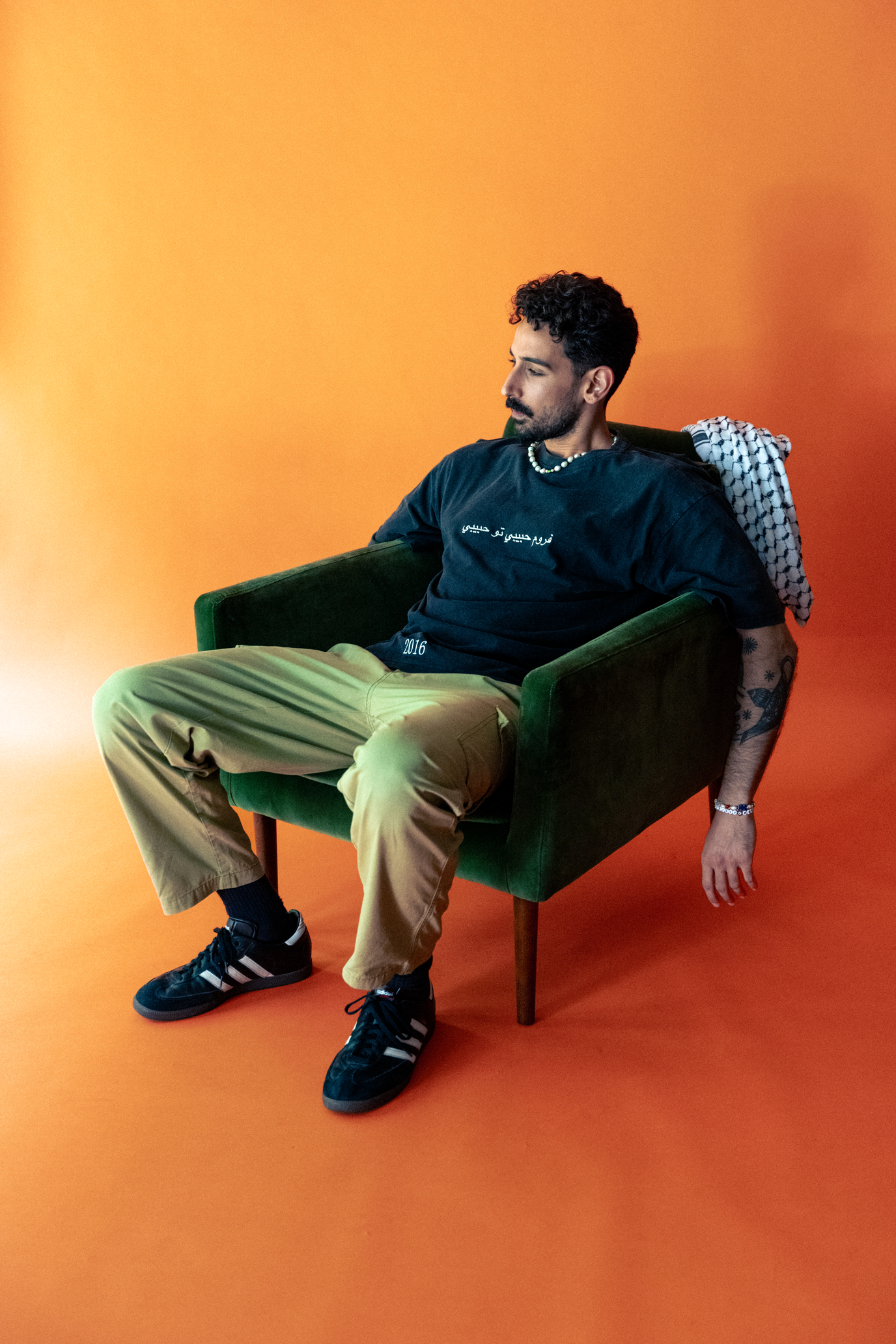 Junge Person sitzt entspannt in einem grünen Sessel vor oranger Hintergrund, trägt schwarze Adidas Schuhe, beige Hosen, schwarze T-Shirt mit weißen Schriftzügen, Perlenkette, Armband, hat Tattoos am Arm und schaut nach unten.