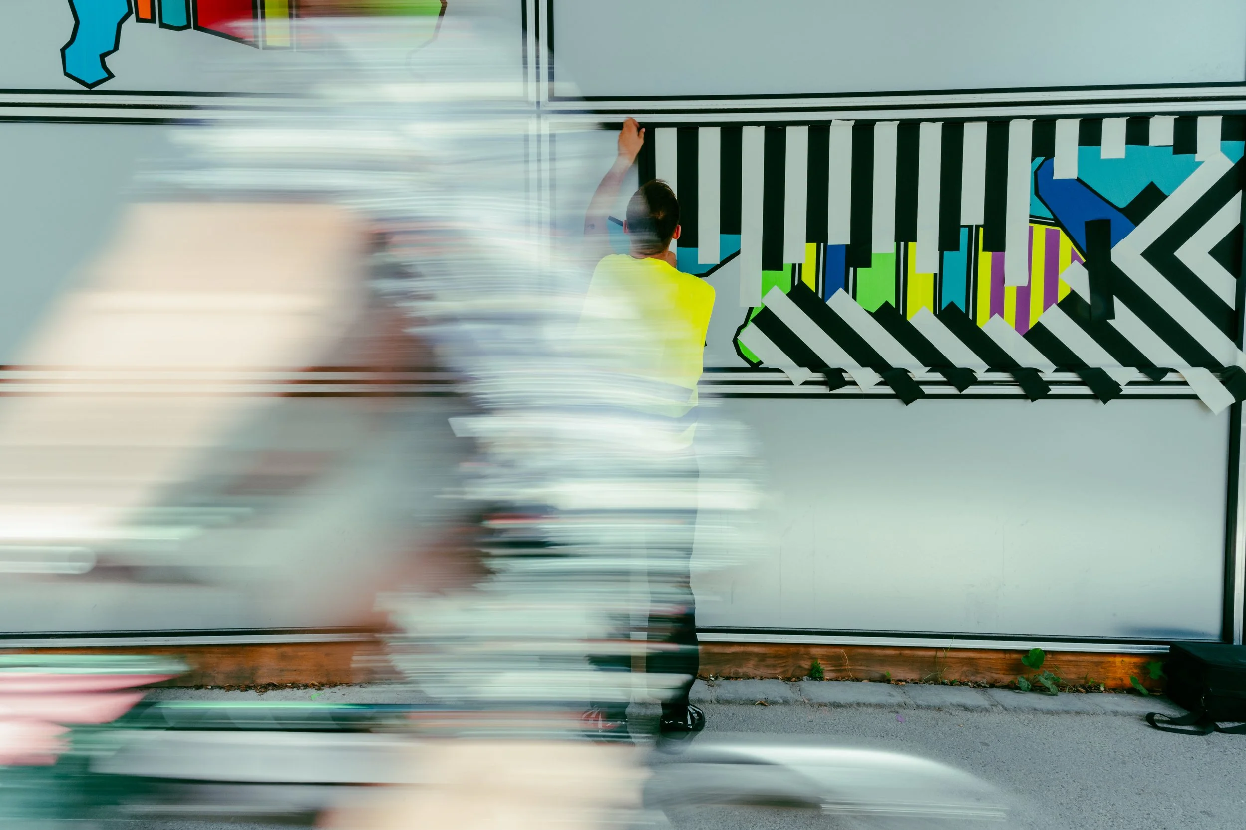 Ein Mann in einem gelben T-Shirt malt an einer Wand, während die Straße an ihm vorbeirauscht.