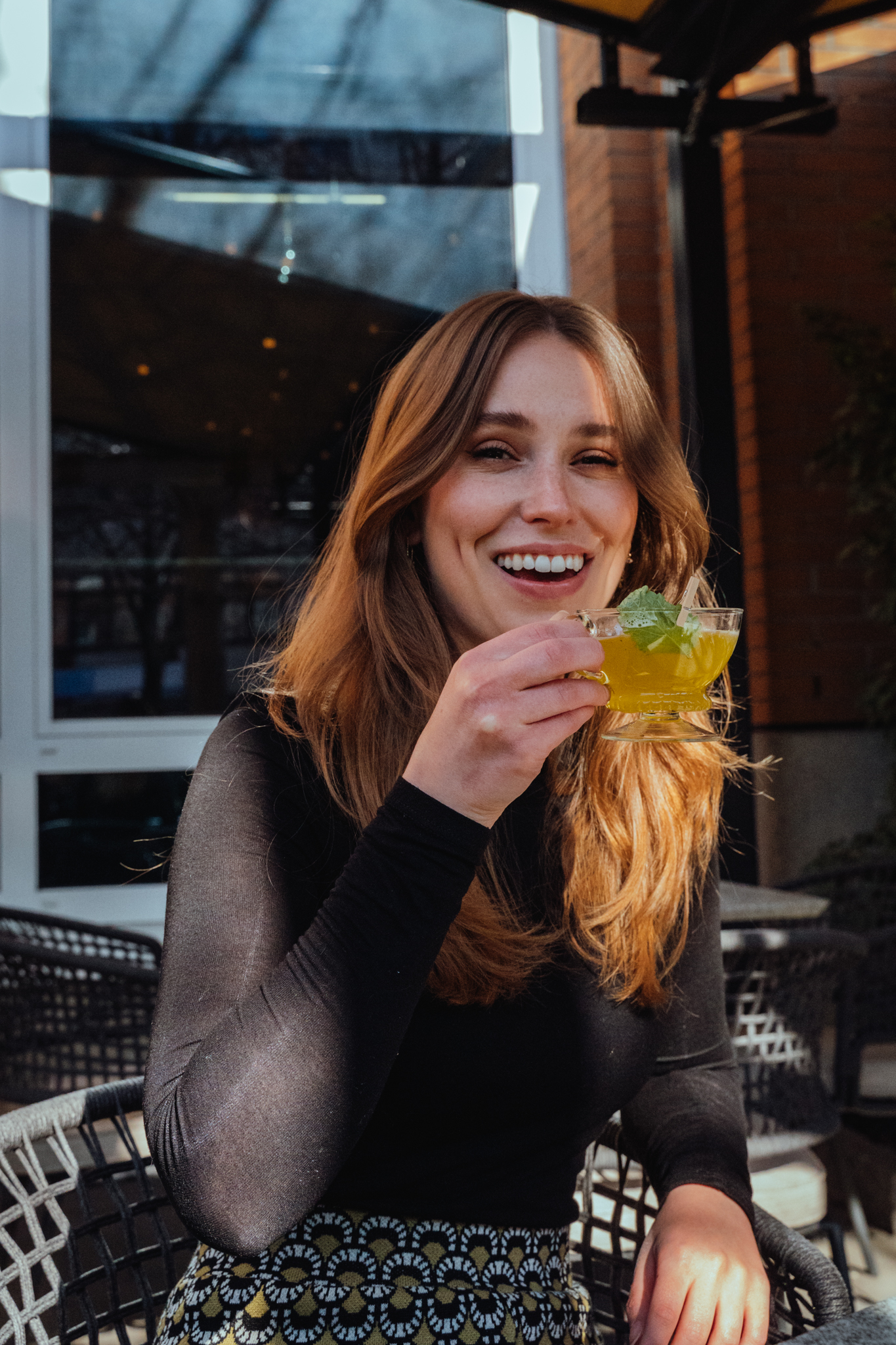 Eine junge Frau sitzt im Freien, hält ein Glas mit gelbem Getränk und grünem Blätter und lächelt.