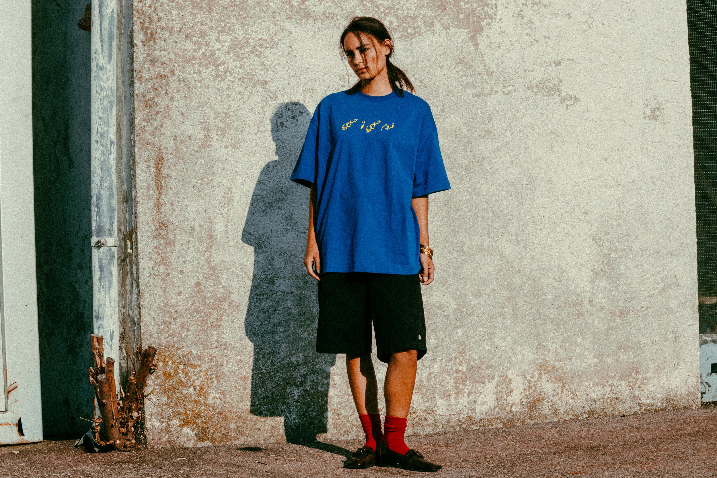 Junge Frau steht an einer Wand mit Schatten. Sie trägt ein blaues T-Shirt, schwarze Shorts, rote Socken und schwarze Schuhe.