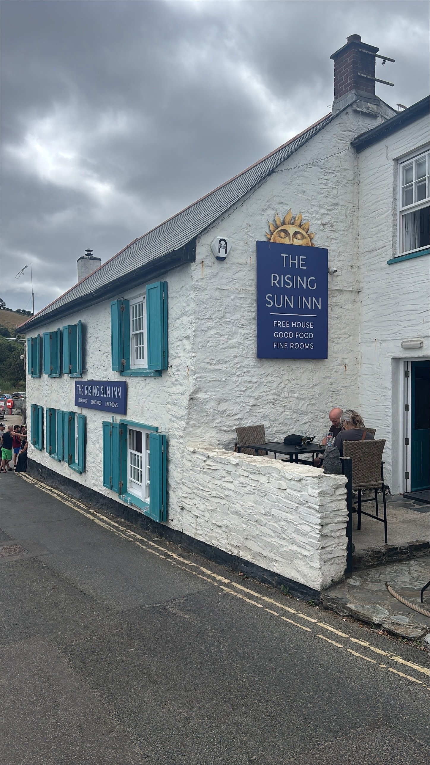 A white building with blue shutters and a blue sign reading 'The Rising Sun Inn,' advertising free house, good food, and fine rooms. A small outdoor seating area with two people sitting and chatting. Overcast sky with dark clouds.