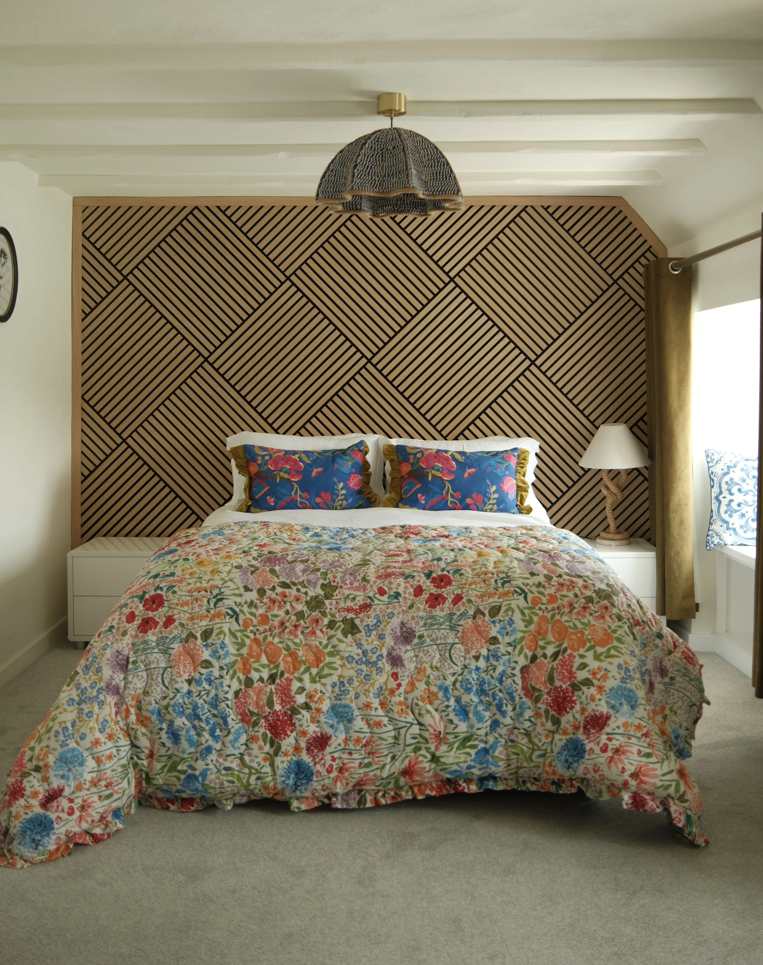 A bedroom with a bed that has a colorful floral bedspread, two pillows with bright floral pillowcases, and a wooden geometric-patterned headboard. There is a white nightstand with a lamp on the right side, a window with a yellow curtain, and a wall-m