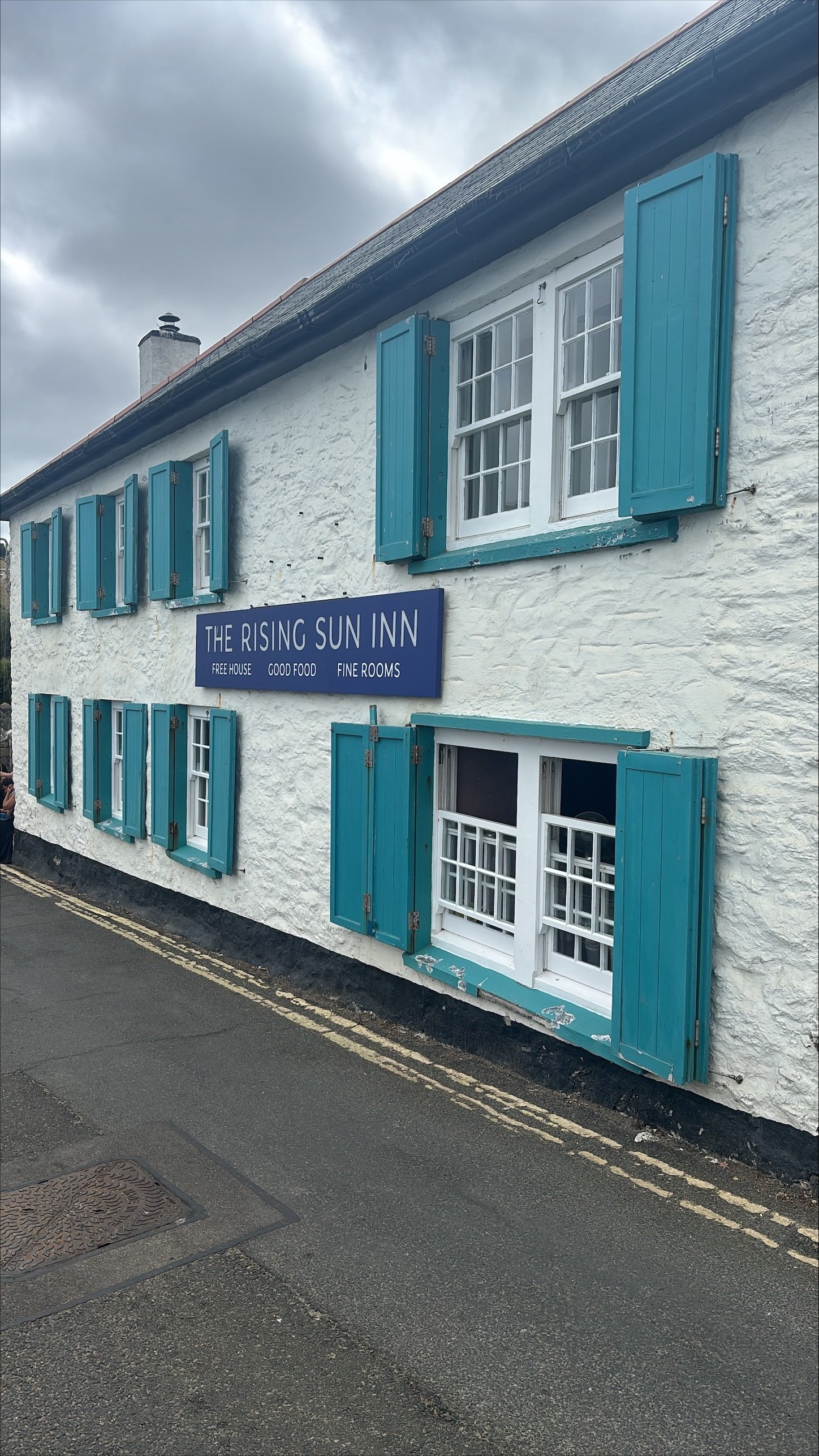 A white building with blue shutters and a sign that reads 'The Rising Sun Inn' offering free house, good food, and fine rooms, situated on a street with asphalt, yellow double lines, and a manhole cover.