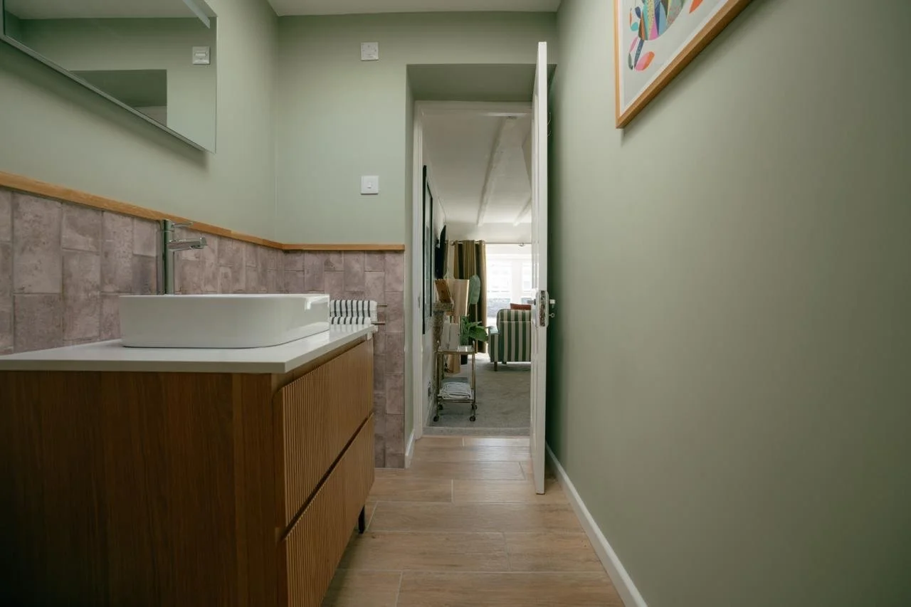 Interior view of a narrow hallway with a wooden dresser and a vessel sink on the left, green walls, and artwork on the right, leading to a living room with striped armchairs and a television in the background.