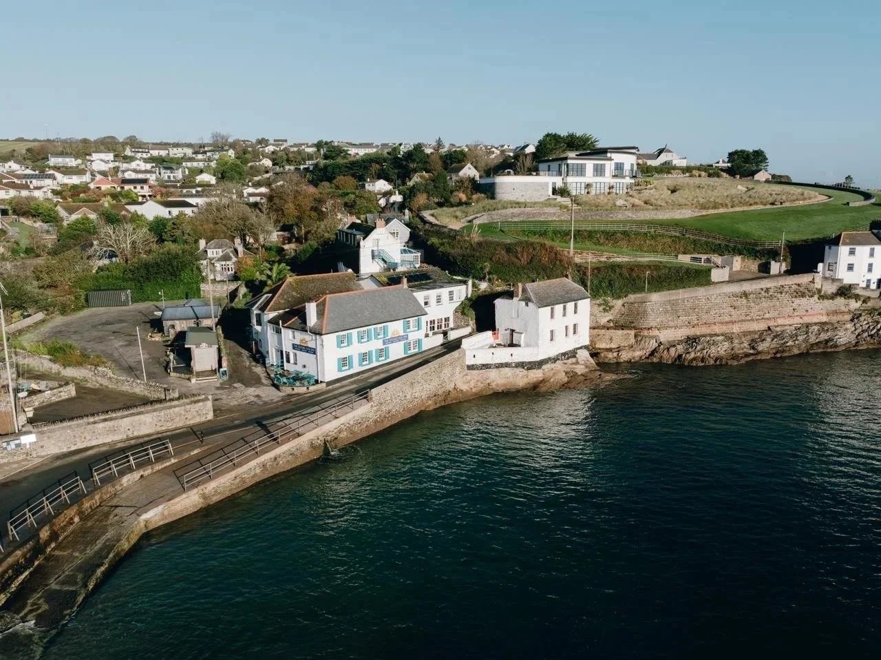 This is a breathtaking photo of the rising sun inn at portmellon, it is a striking white building with blue shutters on the front, the photo is taking by a drone and yu can see the cove, tide is high with blue water, the photo also shows the road.