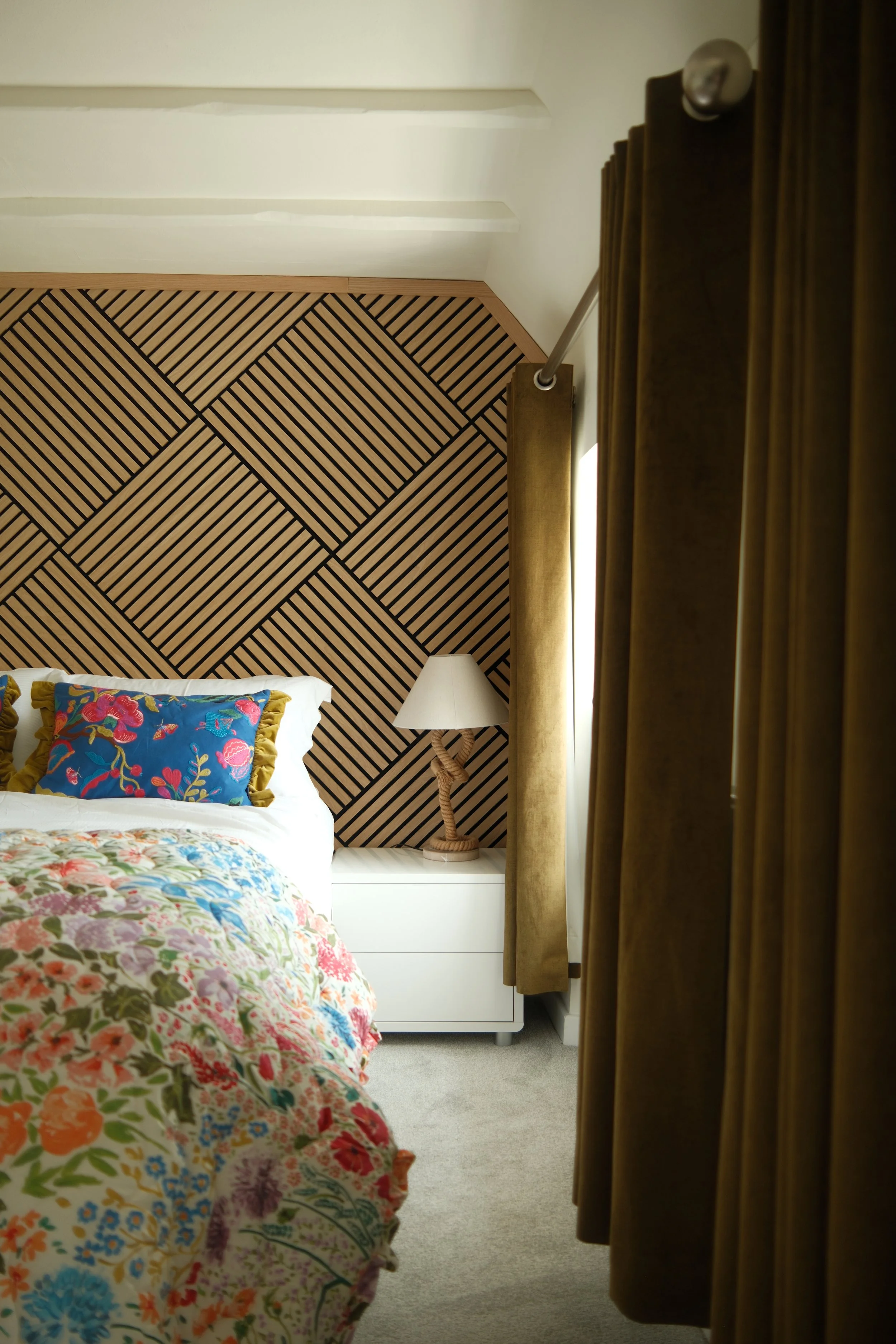 A bedroom with a bed, colorful floral quilt, and a decorative pillow. The headboard is a geometric wooden pattern, and a white nightstand with a lamp is beside the bed. There are brown curtains on the right side.