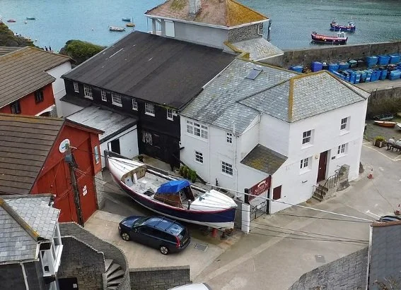 photo shows mevagissey museum which is an old white building two storeys high with a red door and red sign, the back part of the building is black and there is a boat and car next to the entrace, you can also see the sea in the photo with some boat o