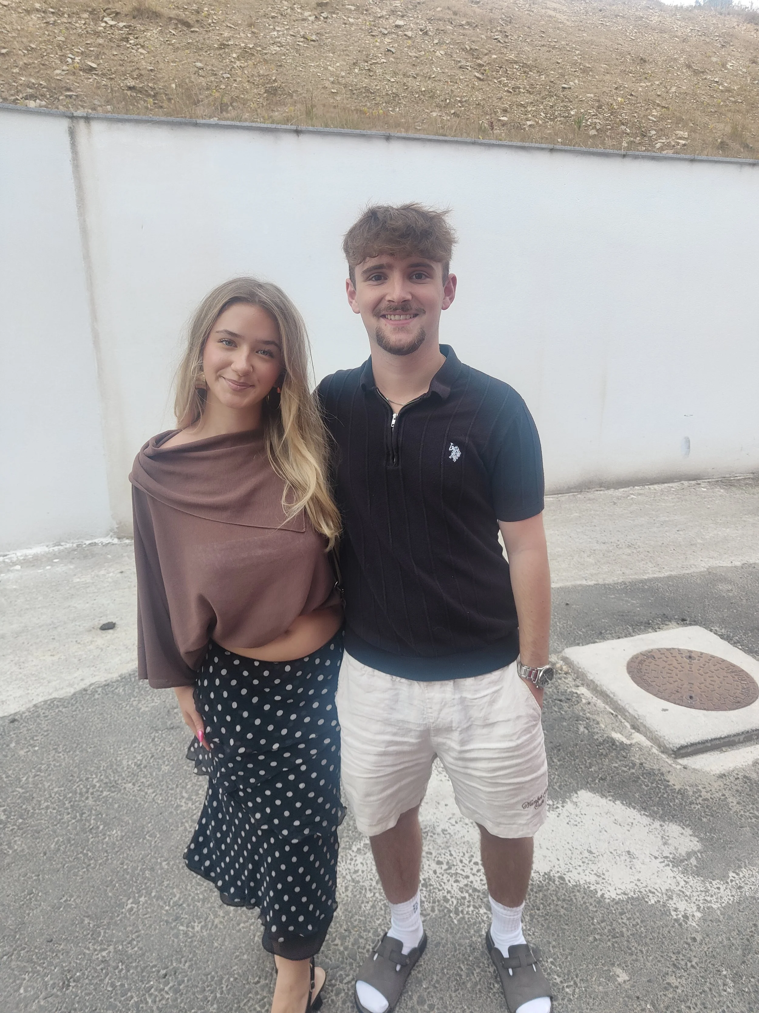 This is a picture of Lotiie and Elliot standing in front of a white wall, Lottie has blonde hair and is wearing a brown top and a balck and white spotted dress, Elliot is wearing a blue top and white shorts, they are both smiling.